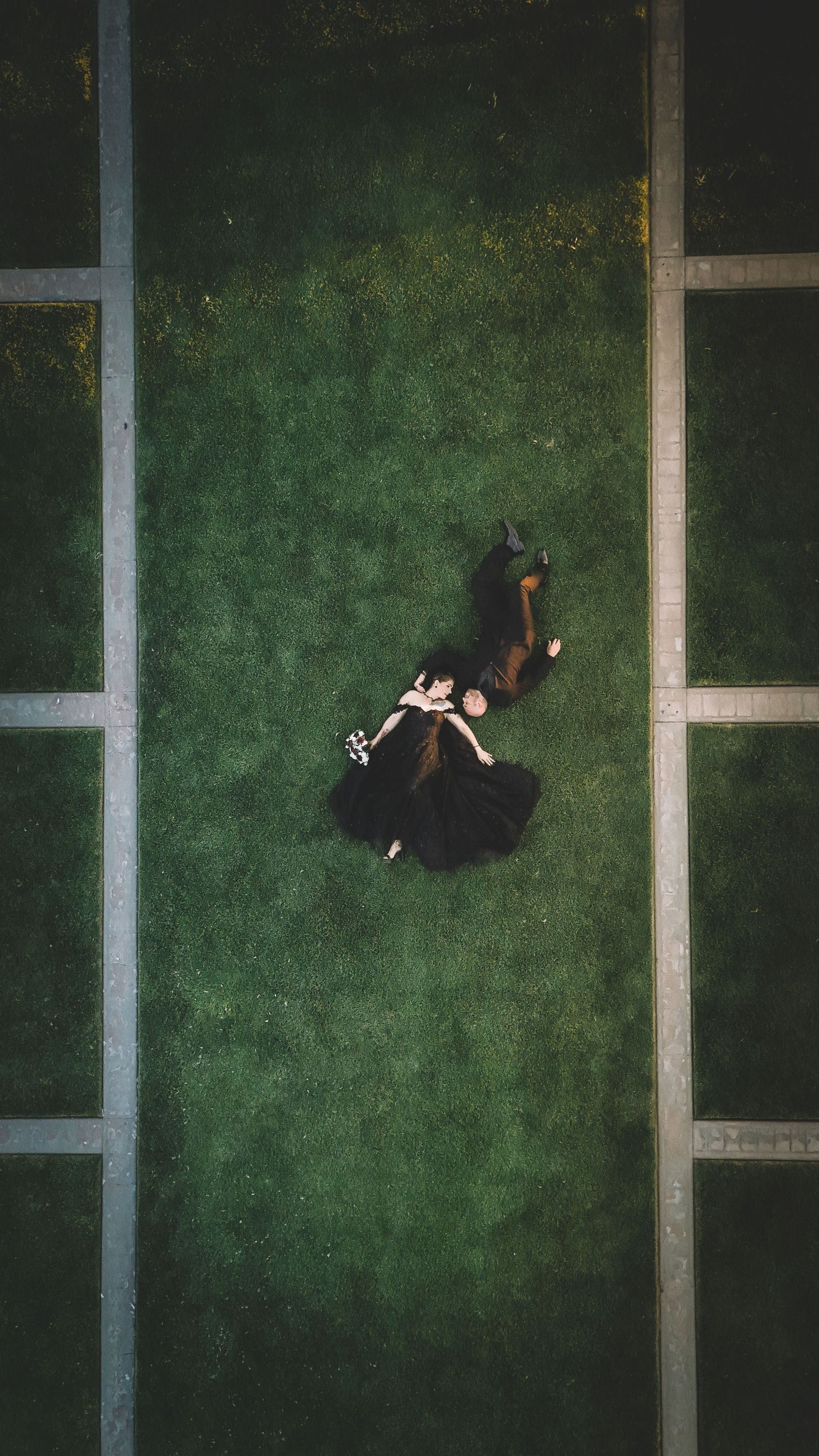 Aerial view of a couple in formal attire lying on a green grass field divided by geometric stone pathways.