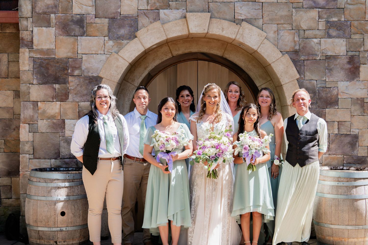 Bride in white gown with bridesmaids in sage green dresses holding floral bouquets posing before stone arch at winery wedding.