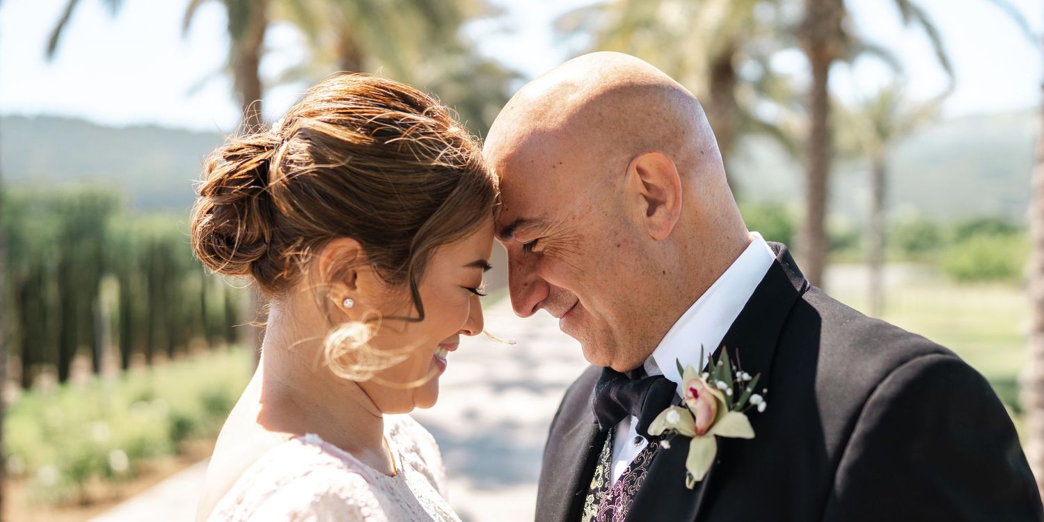 Bride and groom touching foreheads tenderly at an outdoor wedding venue surrounded by palm trees.