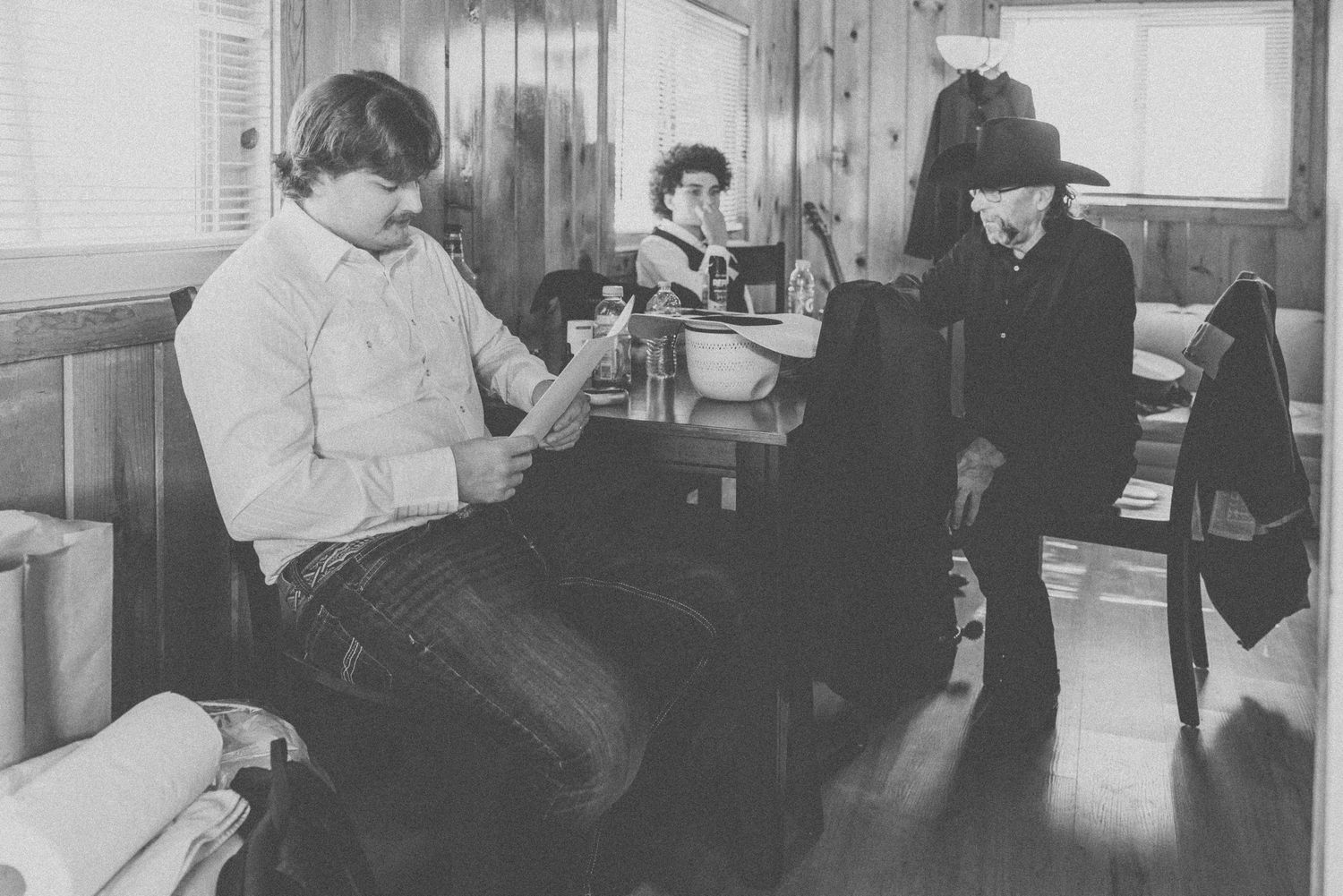 Black and white photo of three people in a rustic room, one seated reading, another at a sink, and one standing in a hat.