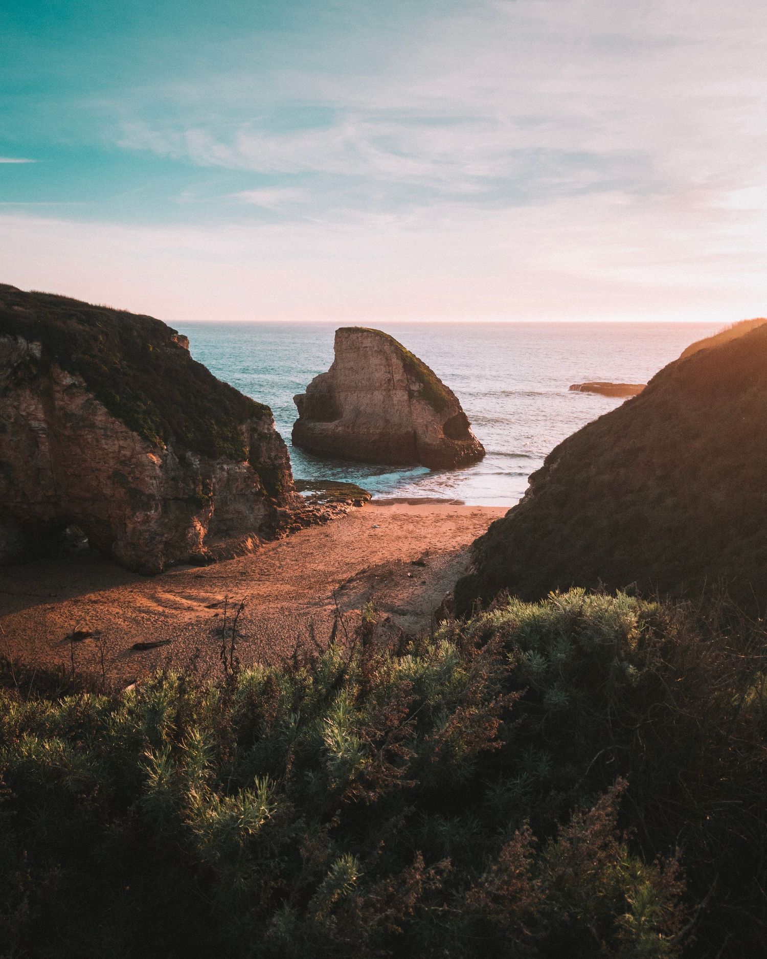 Scenic coastal cove at sunset with rugged cliffs, sandy beach, and a large sea rock framed by lush green vegetation.