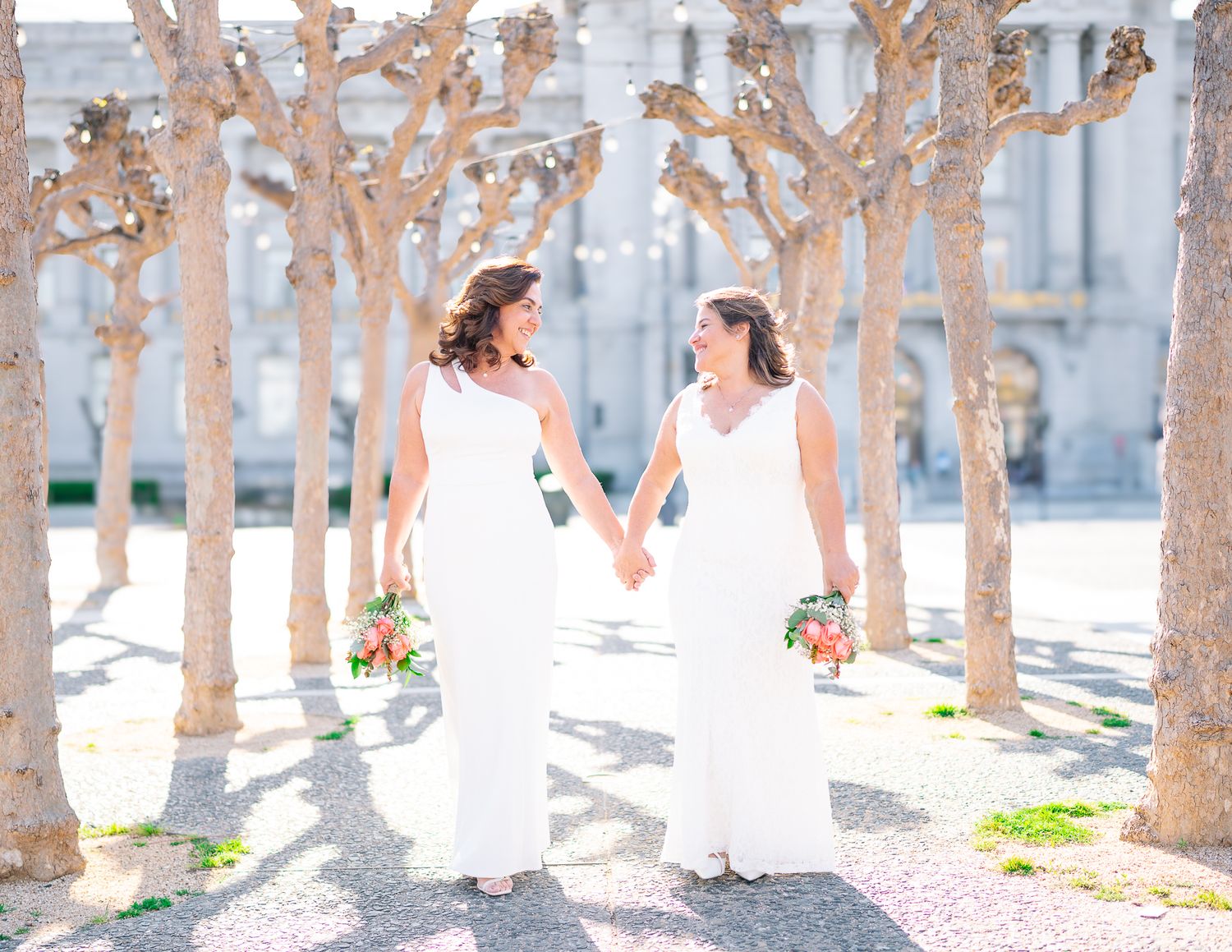 Two brides in white dresses hold hands and bouquets while walking among trees at a sunny outdoor wedding.