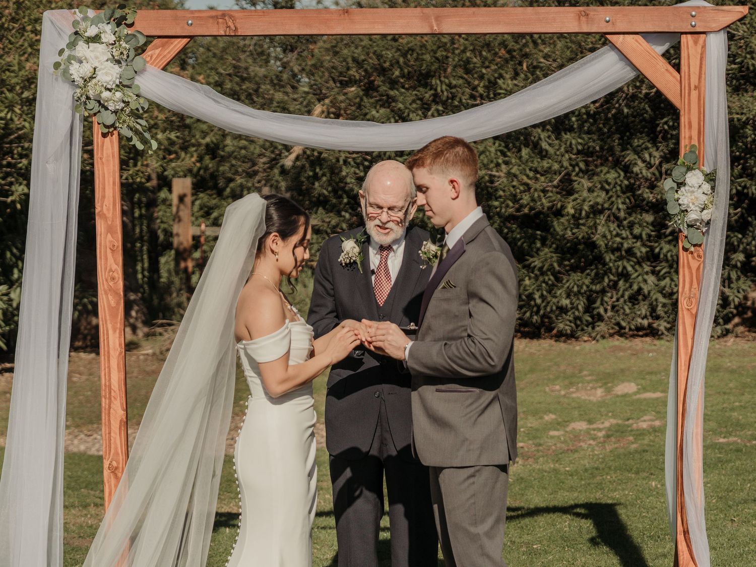 Bride and groom exchanging rings at outdoor wedding ceremony under rustic wooden arch with floral decorations.