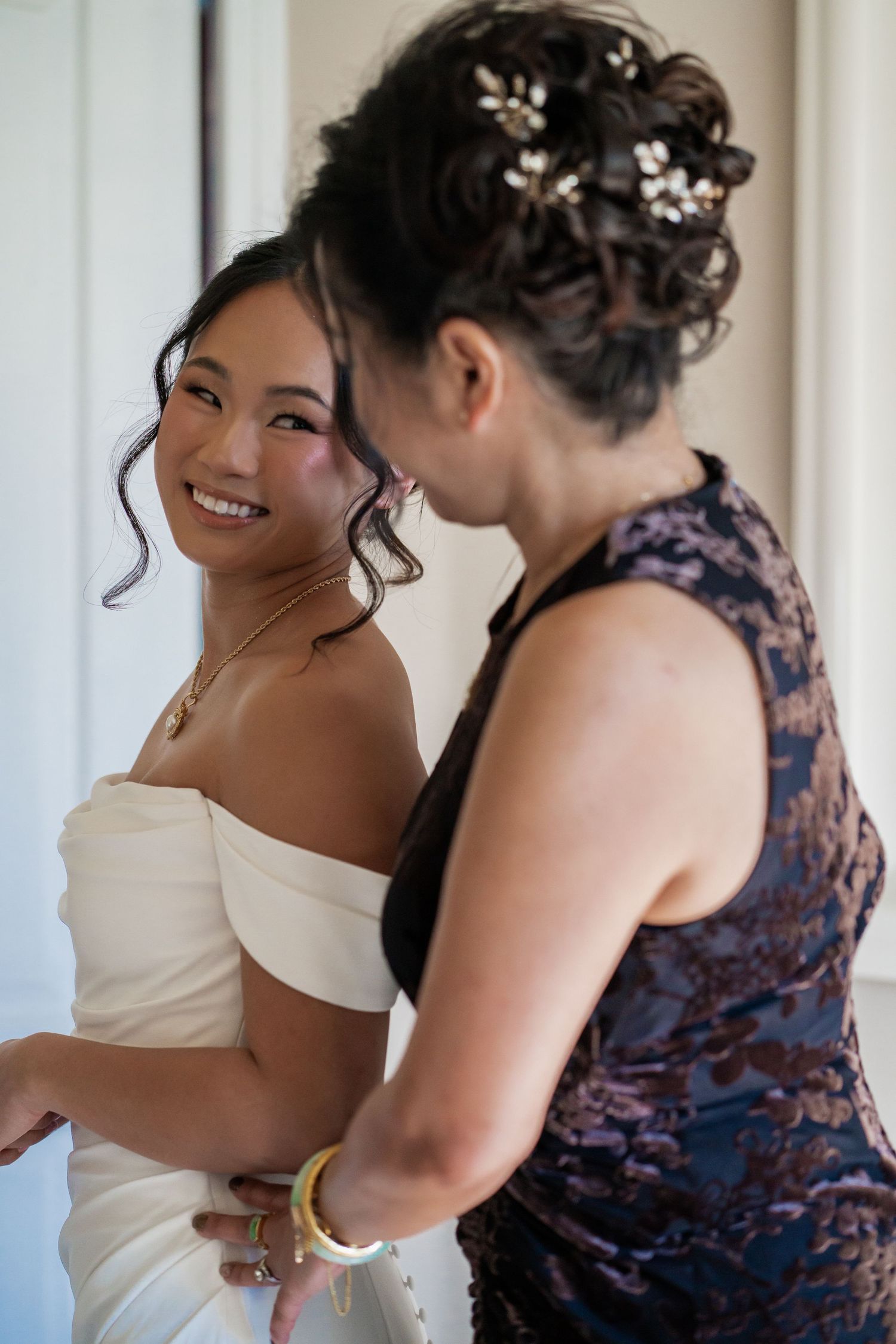 A smiling bride in a white dress gets help from a woman in a floral dress, with floral hair accessories.