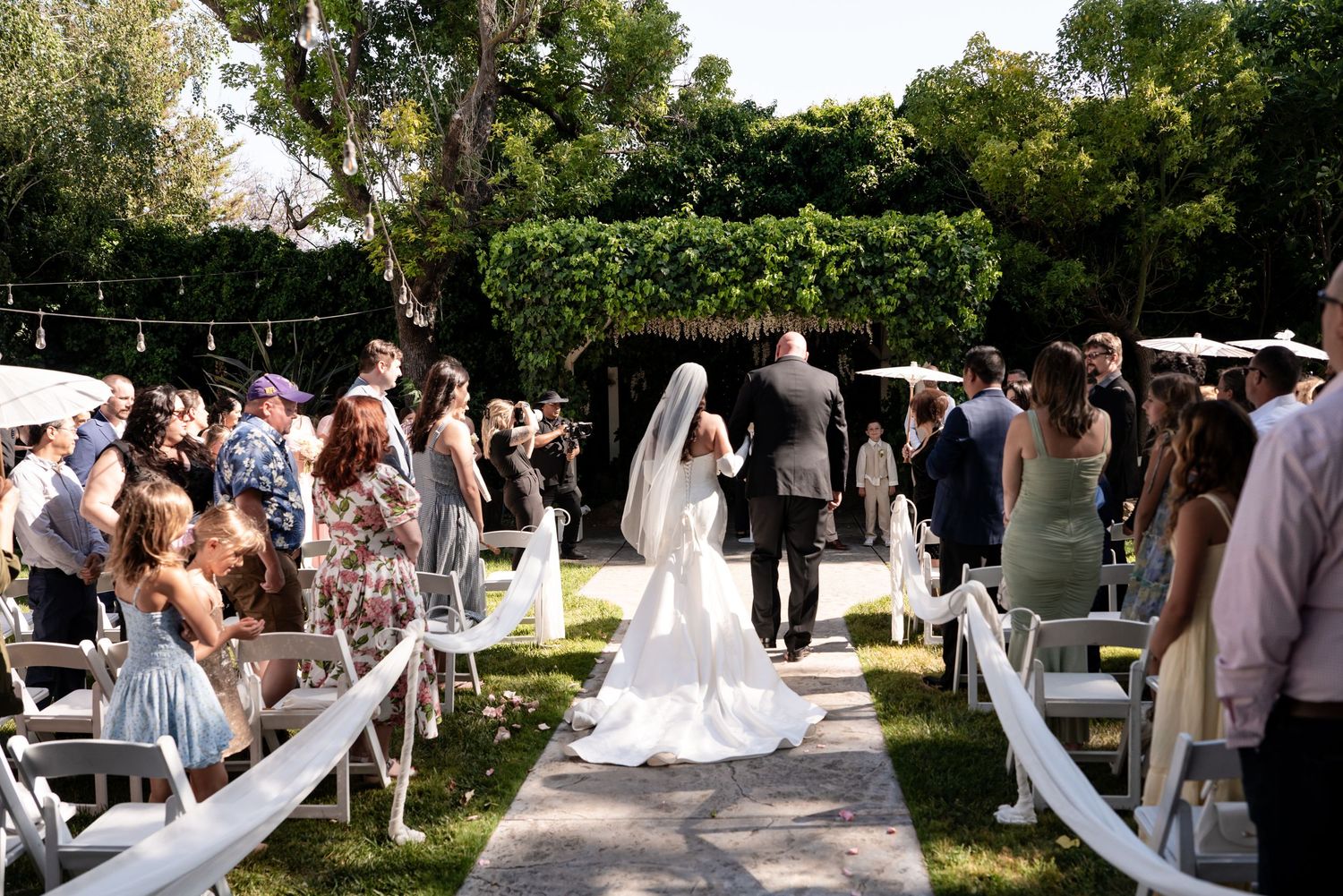 Bride walks down outdoor wedding aisle in white gown and veil, surrounded by seated guests under string lights.