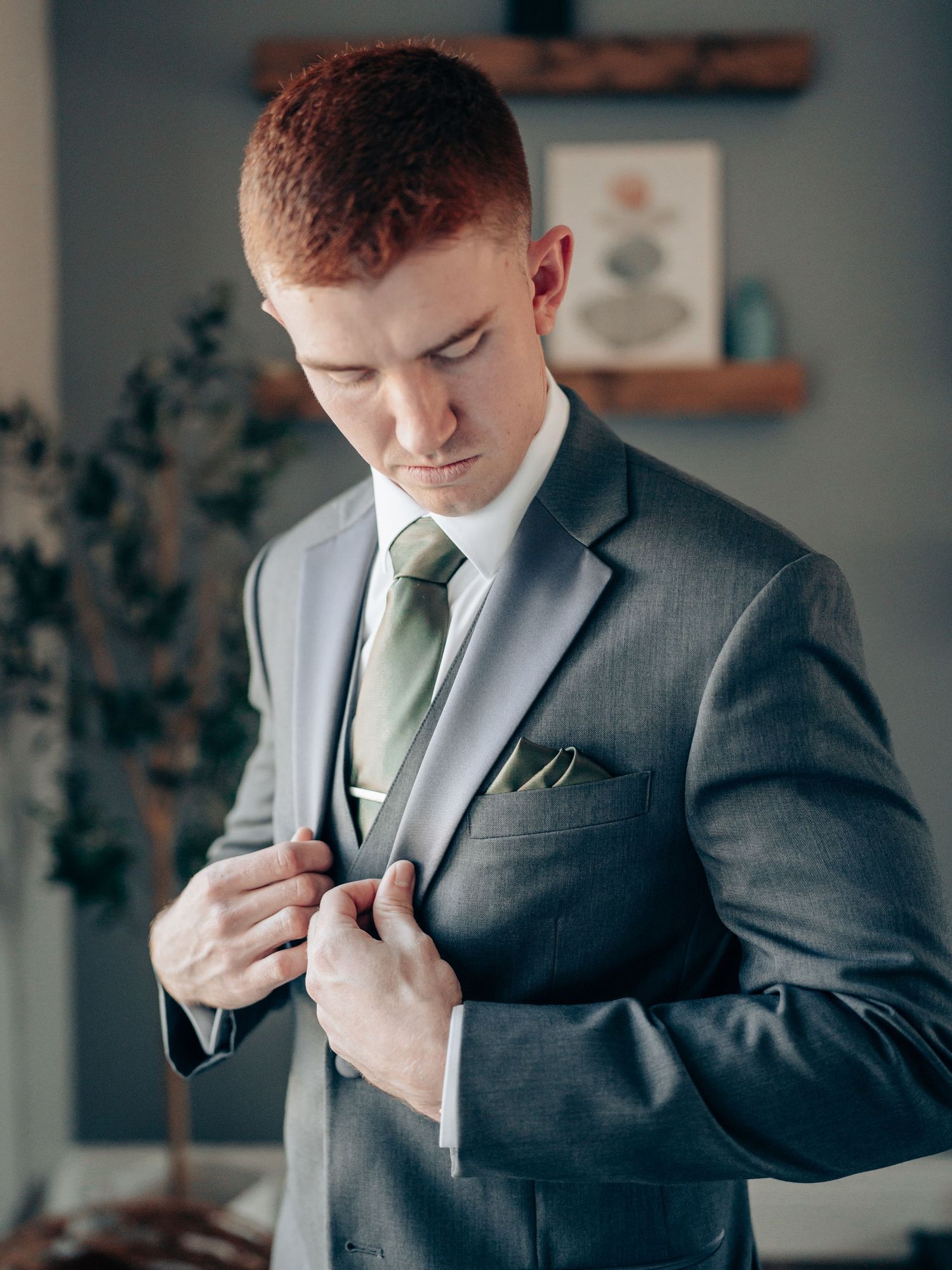 A young man in a gray suit with green tie and pocket square adjusts his jacket button indoors.