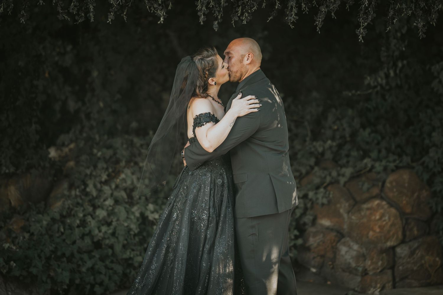Couple sharing a romantic kiss in wedding attire, bride in black gown with veil, groom in dark suit, ivy backdrop.