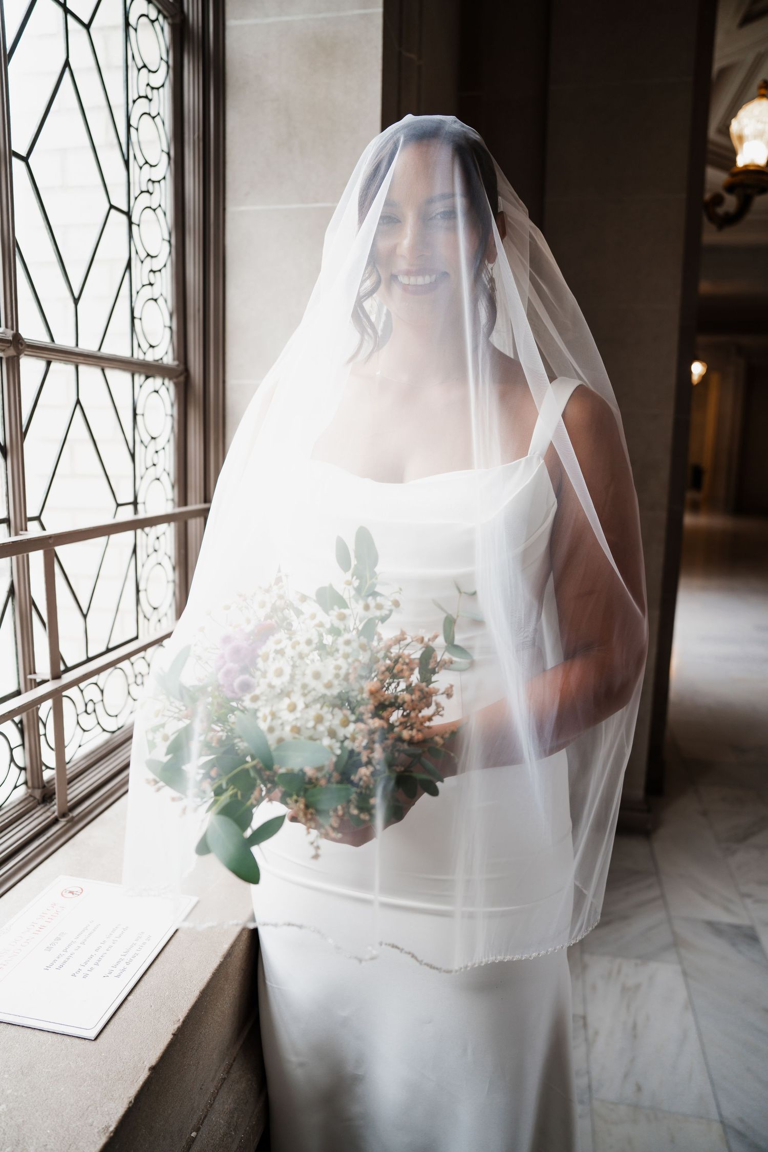 Bride in white wedding dress and veil holds delicate bouquet while standing by ornate window.