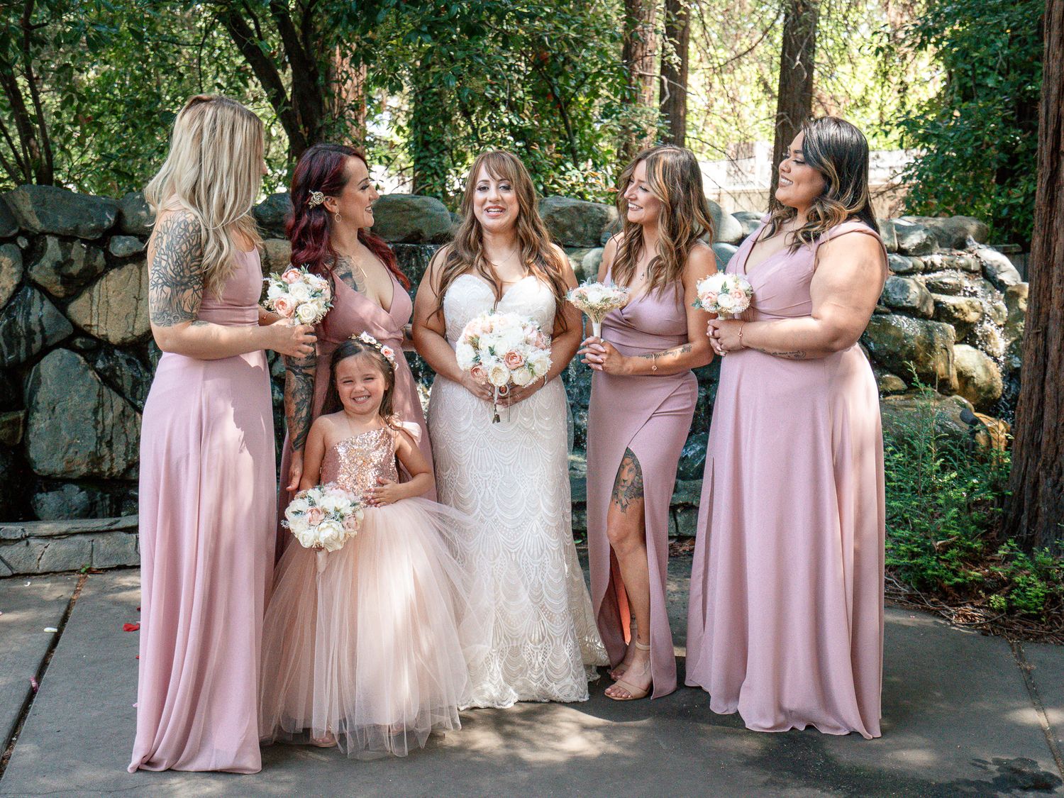 Bride in white gown poses with four bridesmaids and flower girl in blush pink dresses outdoors.