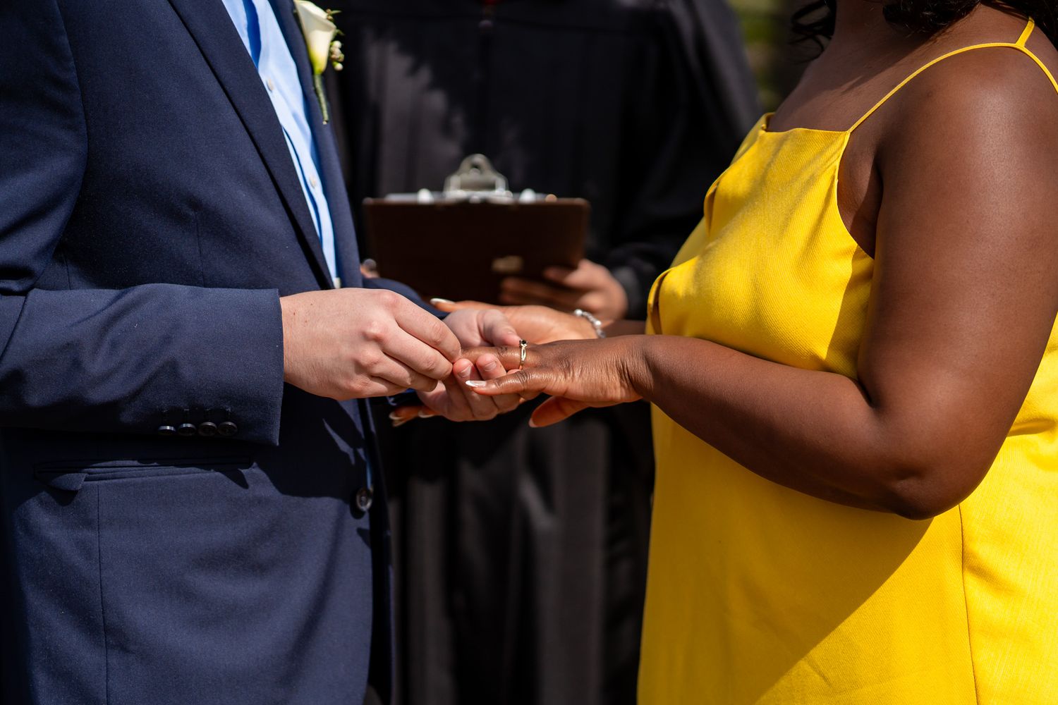 Groom places wedding ring on brides finger during outdoor ceremony, bride wearing yellow dress.