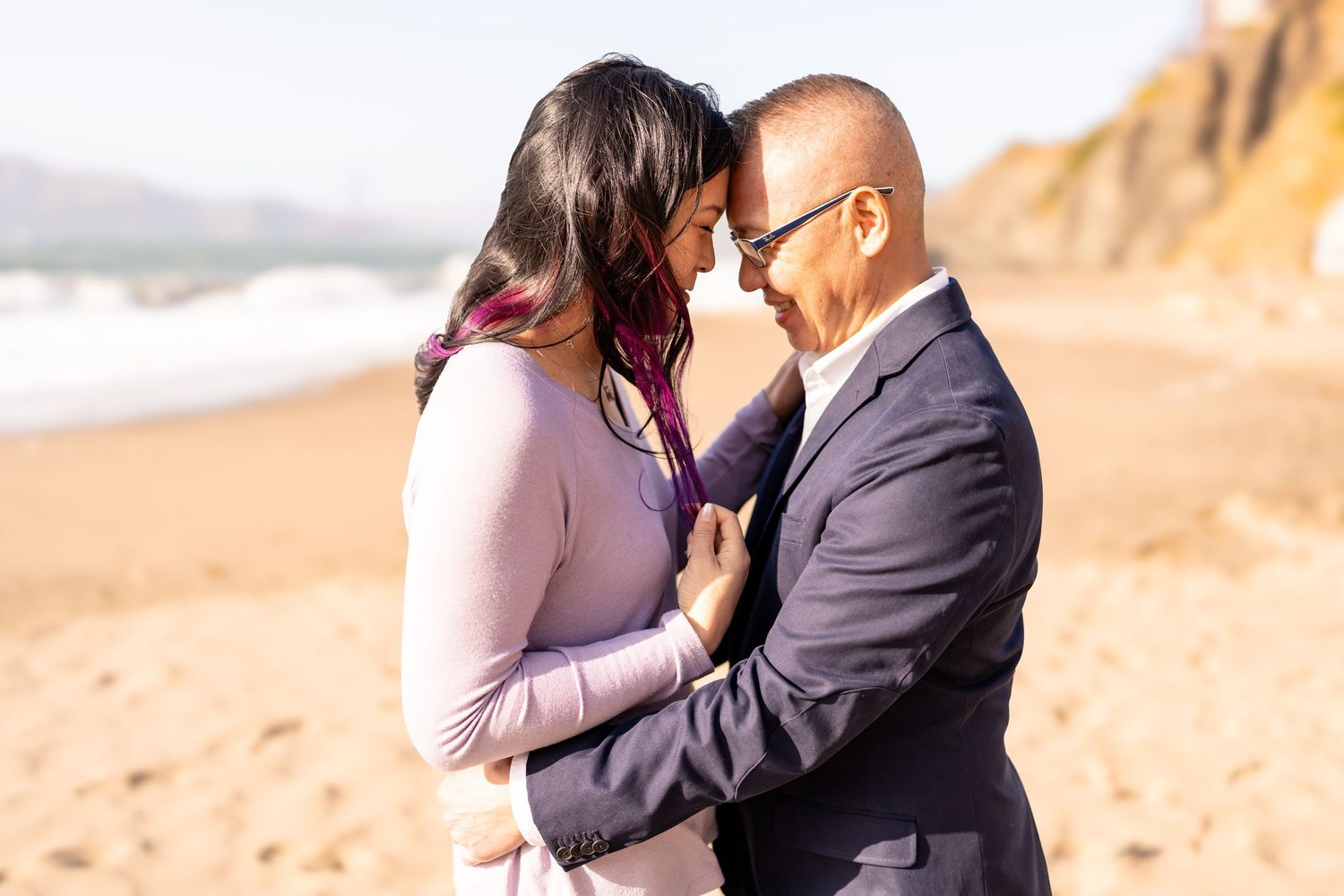 Romantic couple embraces on a beach at sunset with rocky coastline in the background.