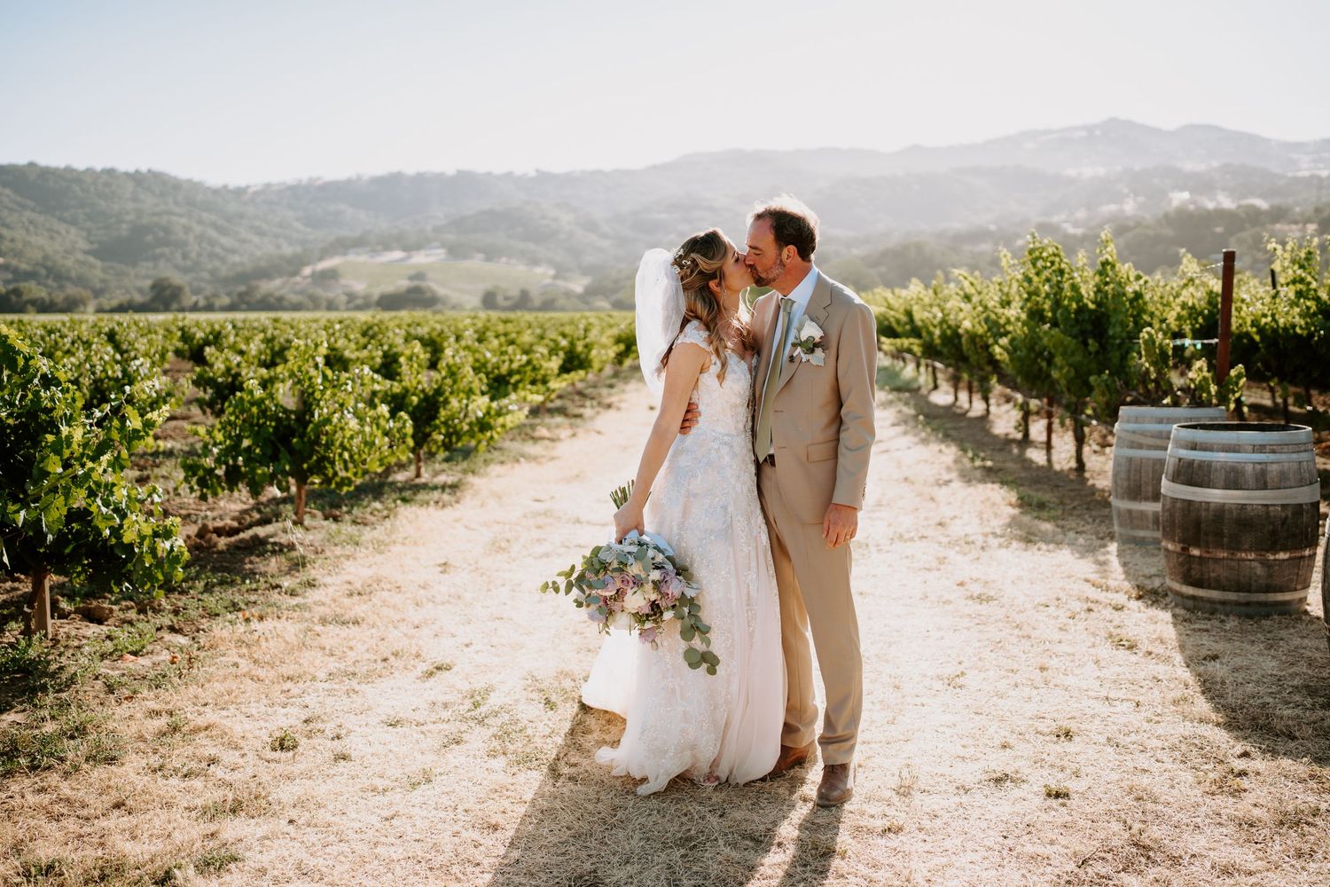 Wedding couple walking through scenic vineyard with mountain backdrop.