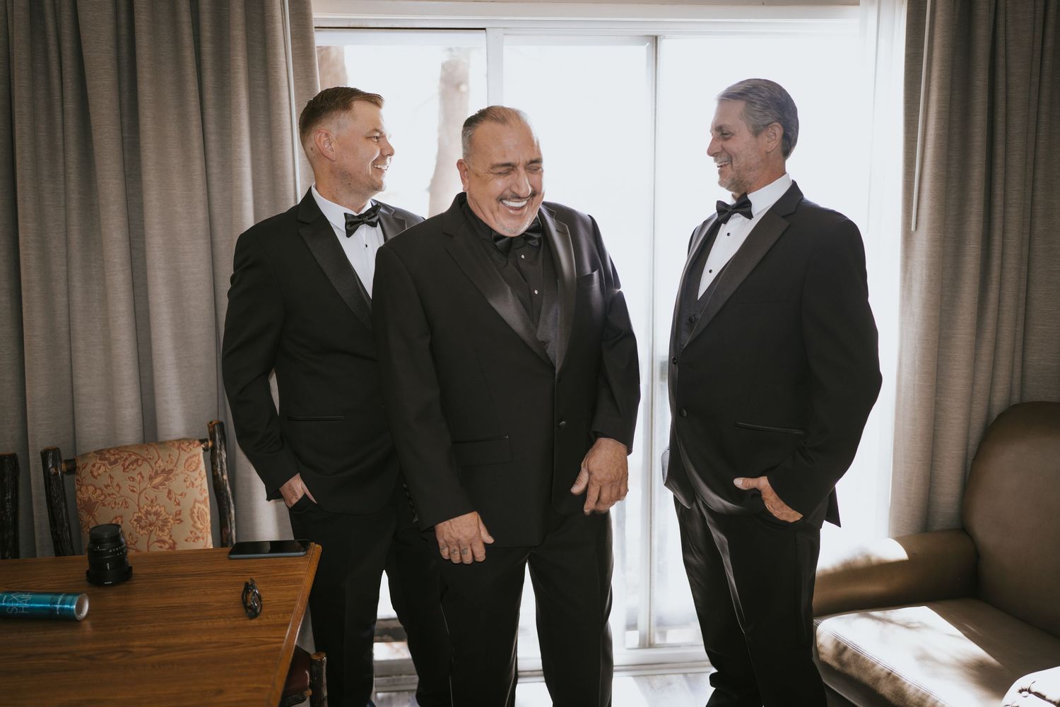 Three men in black tuxedos and bow ties laughing together in a bright room during a wedding celebration.