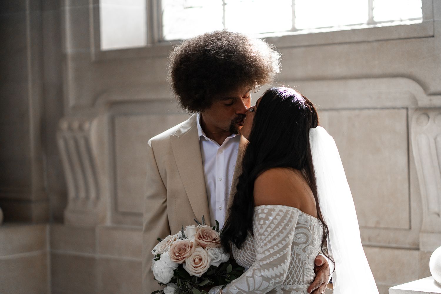 Newlywed couple sharing an intimate moment with white roses bouquet in elegant indoor wedding venue.