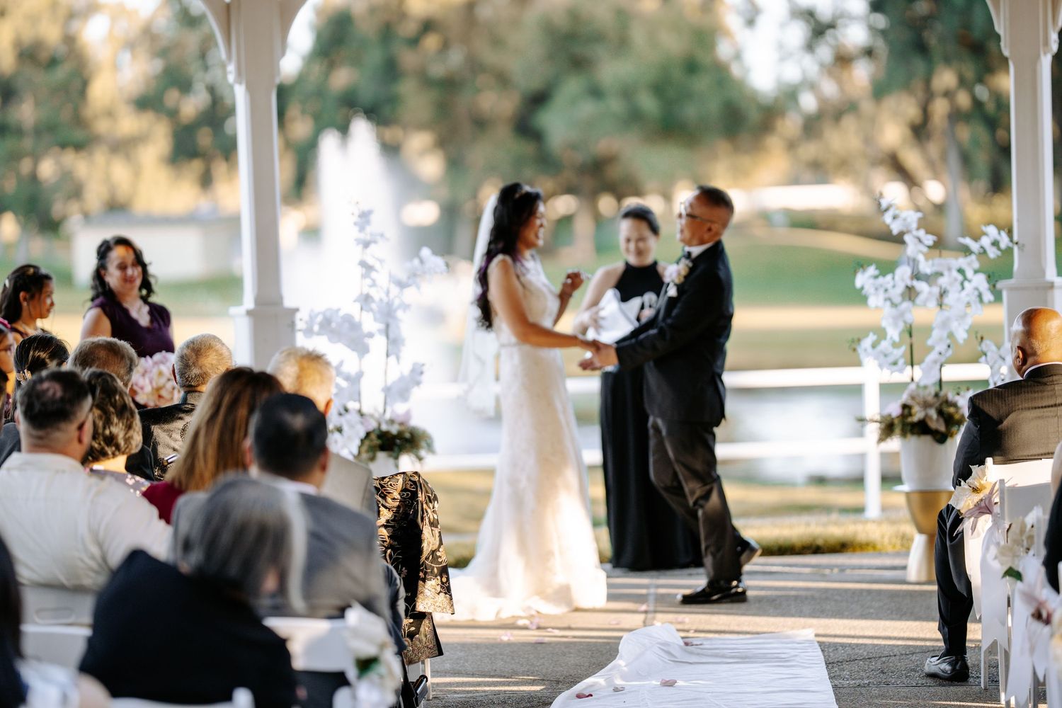 Outdoor wedding ceremony under a white gazebo with bride and groom exchanging vows, guests seated, and a scenic lake backdrop.