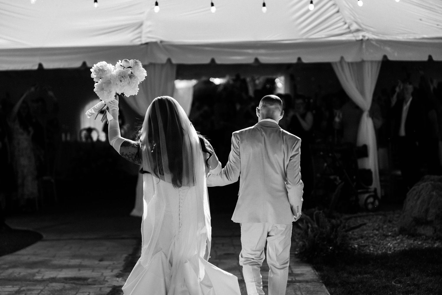 Black and white photo of a bride and groom walking hand in hand toward a tented wedding reception venue.