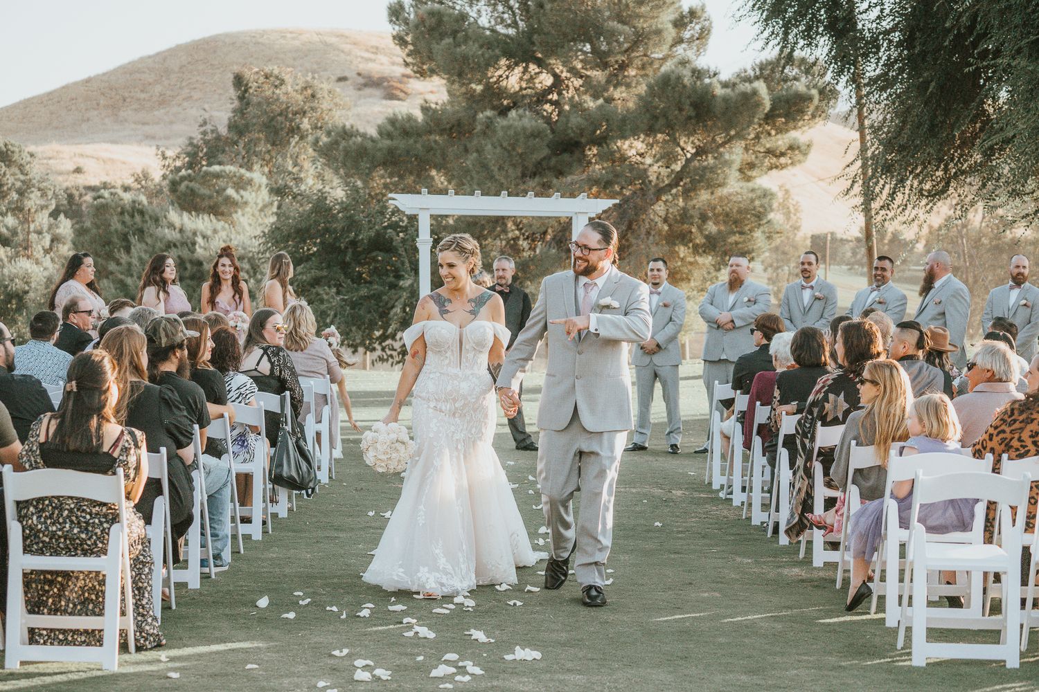Bride and groom walk down the aisle at an outdoor wedding ceremony with guests seated on white chairs.