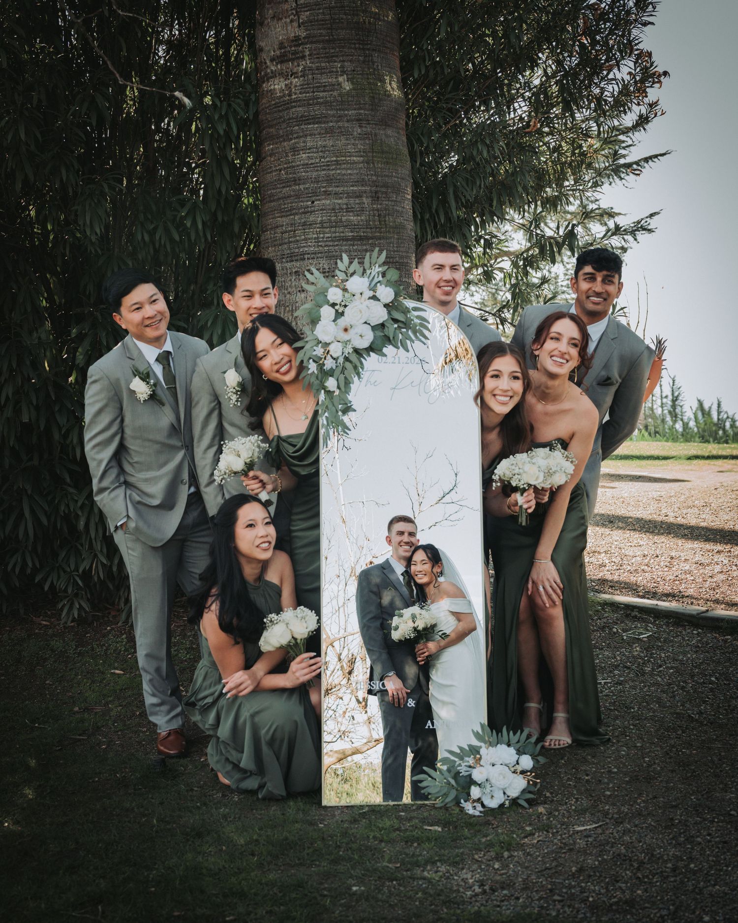 Wedding party in sage green attire posing around a floral portrait sign near a tree at outdoor wedding venue.