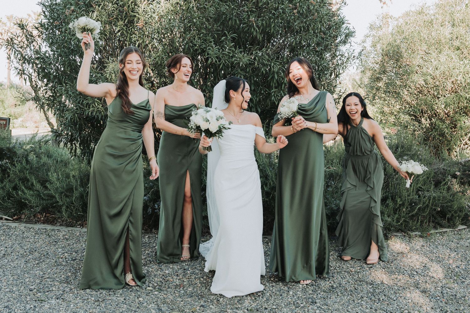 Bride in white gown with four bridesmaids in olive green dresses celebrating outdoors.