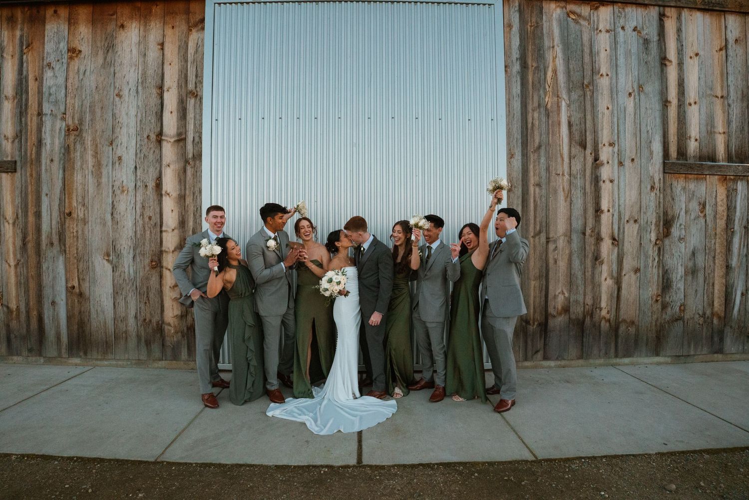 Wedding party in sage green and gray attire celebrating outside rustic barn venue.