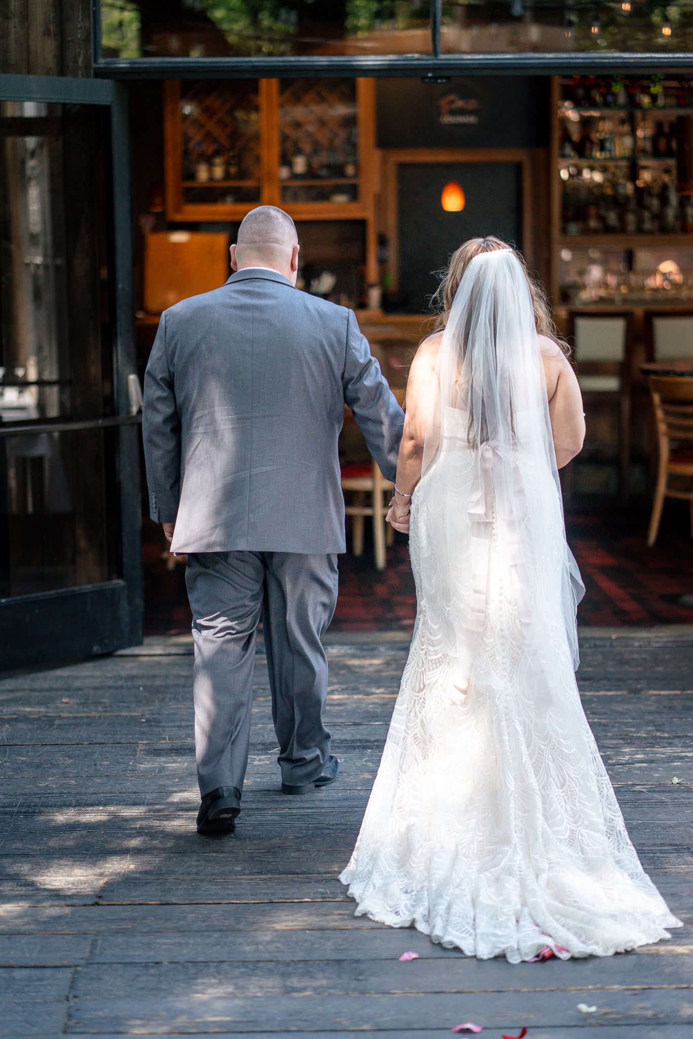 Bride in white lace gown and veil walks hand-in-hand with groom in gray suit toward restaurant entrance.