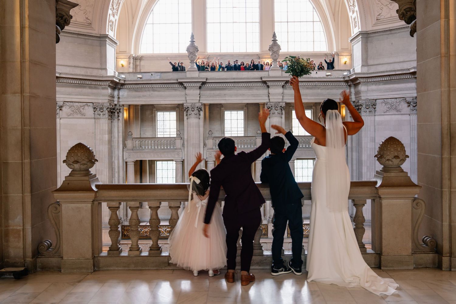 Wedding party celebrating with raised arms in grand historic building interior.
