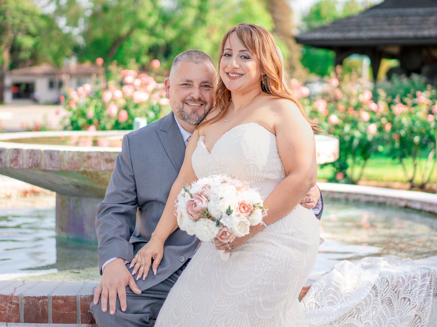 Smiling bride and groom pose together by a fountain in a lush garden wedding setting.