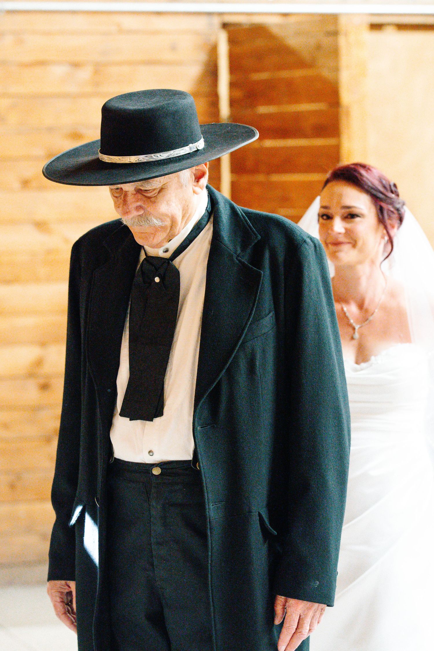 Elderly man in black wide-brim hat and coat walks ahead of smiling bride in white veil at rustic barn wedding.