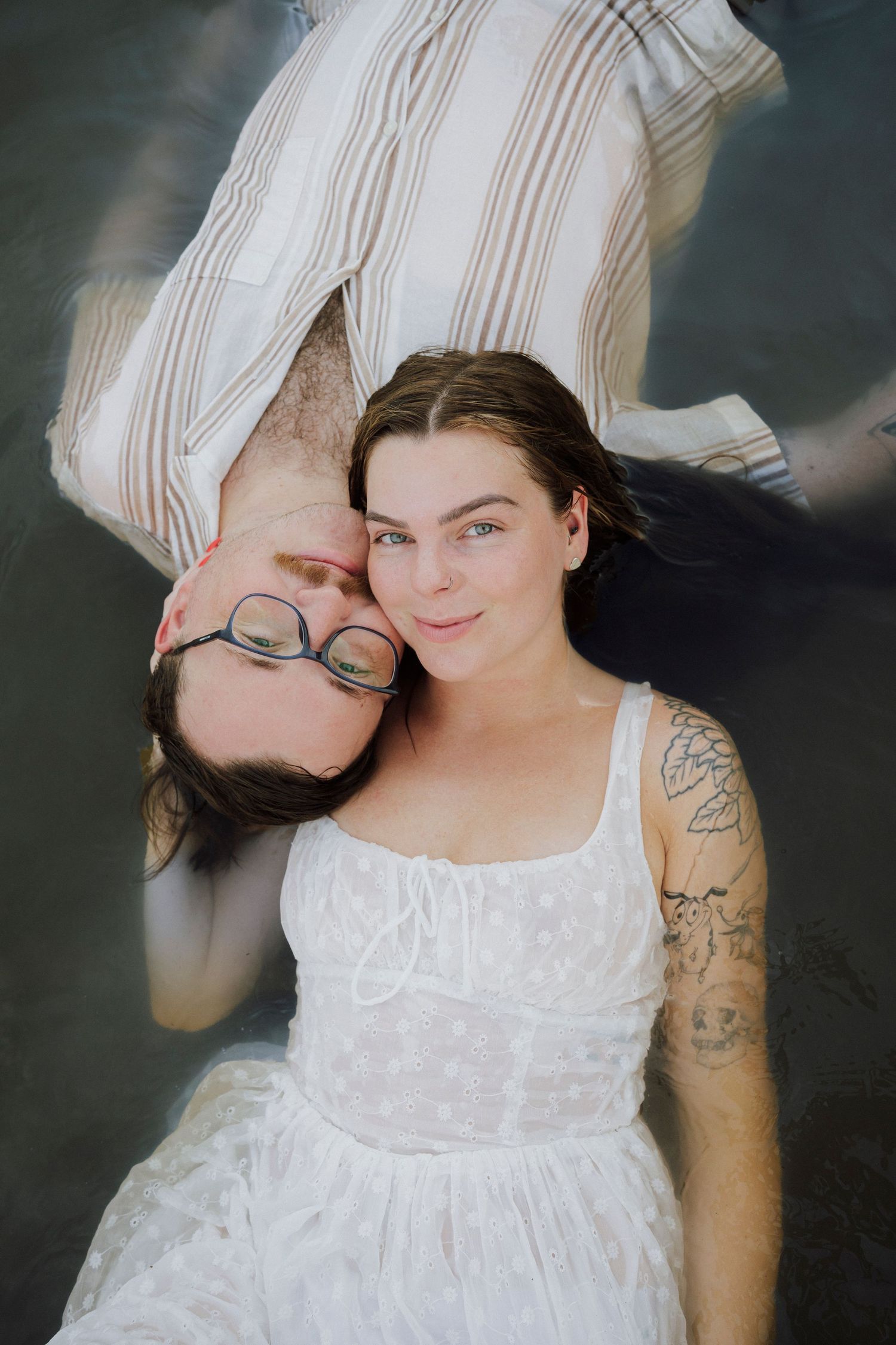 Two people lying down together, one in a white dress with tattoos smiling at camera, other in striped shirt wearing glasses.