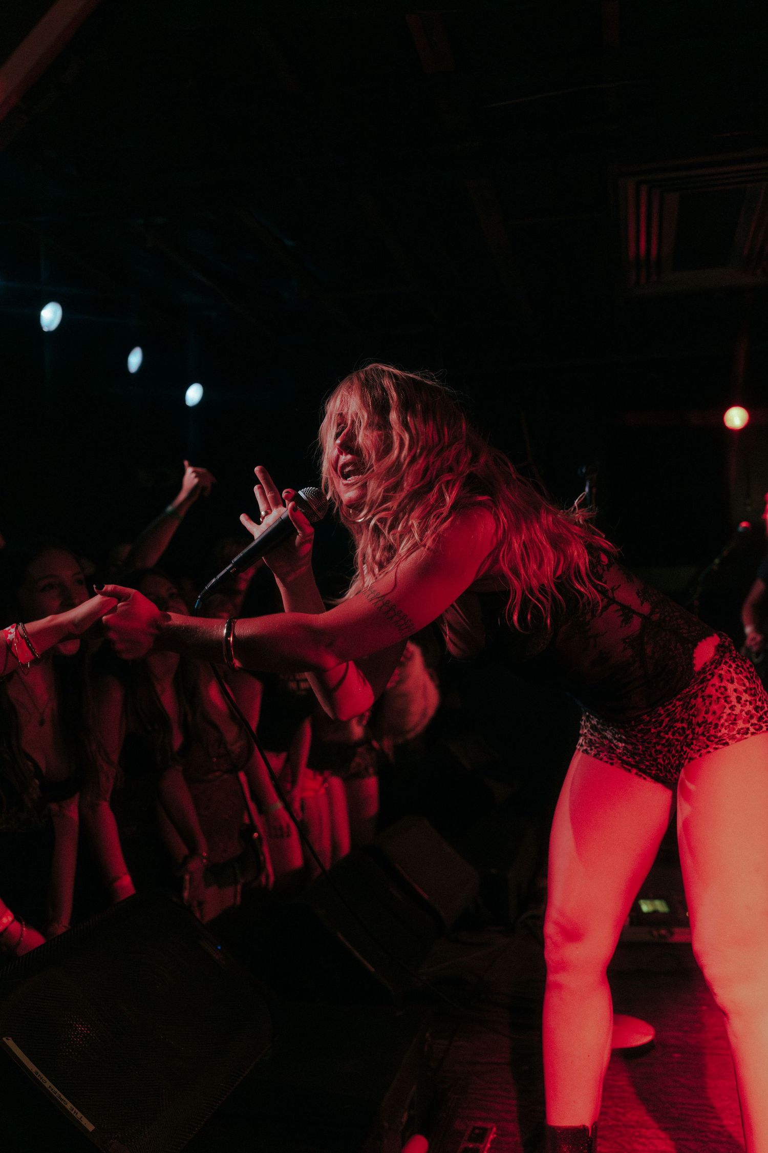 Female singer performing on stage, leaning toward crowd with microphone under red lights at a live concert.