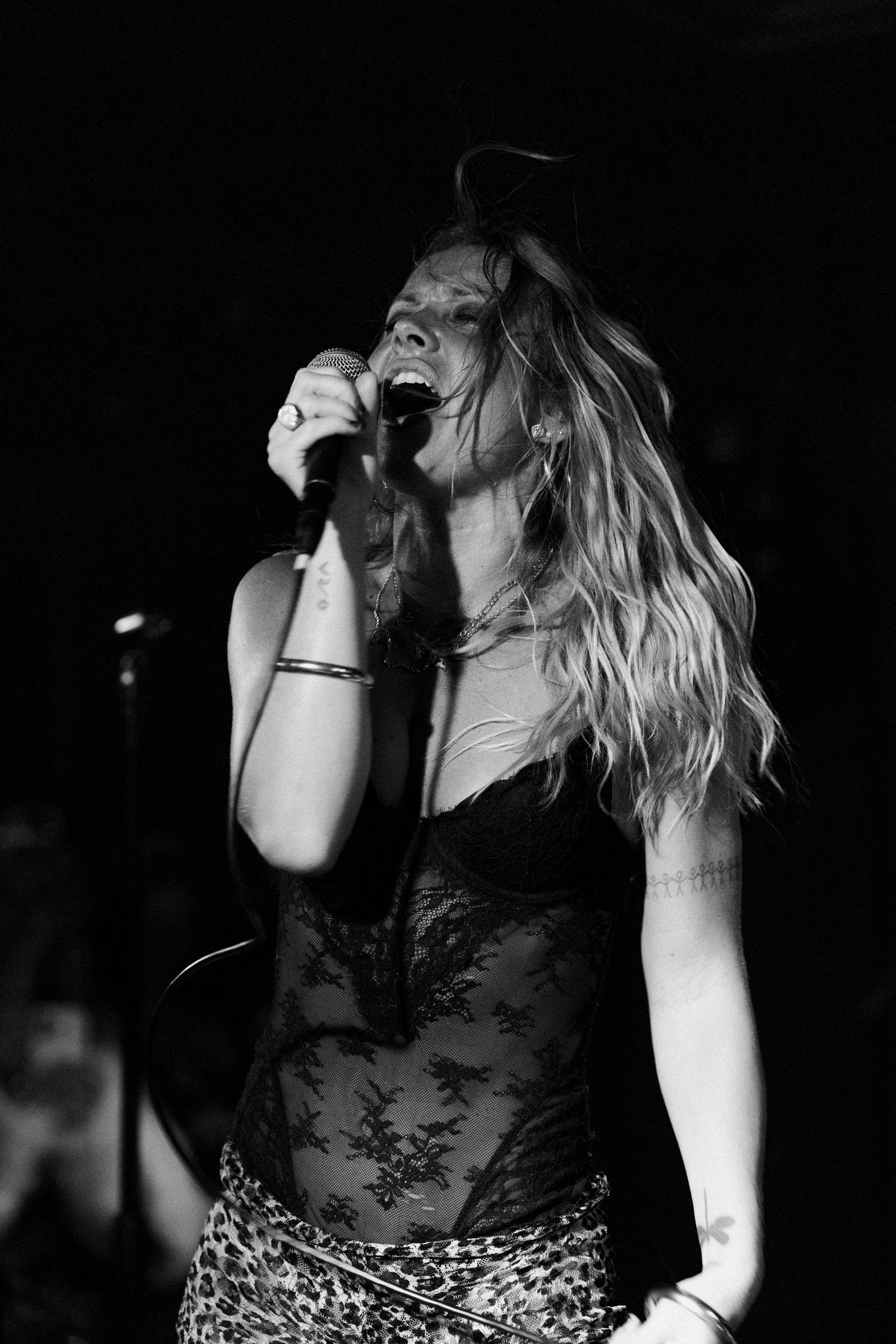 Black and white photo of a female singer performing on stage, holding a microphone, wearing a lace top and leopard skirt.