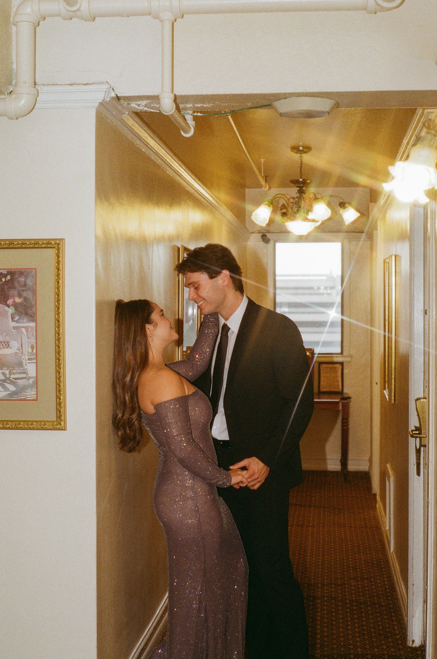 Couple in formal attire sharing a romantic moment in a warmly lit hotel hallway.