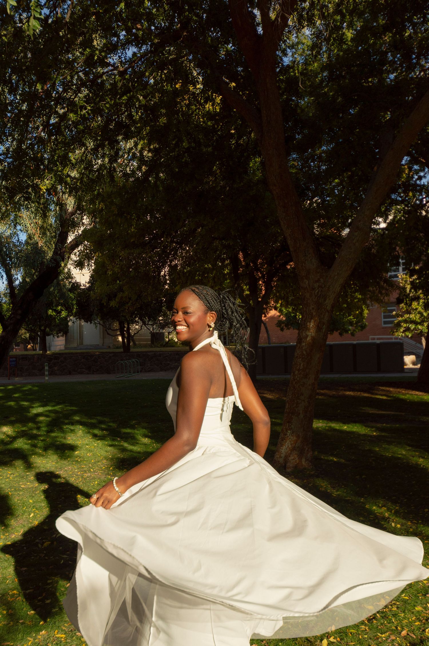 Young Black woman in white halter dress twirling under trees in sunny outdoor park setting.