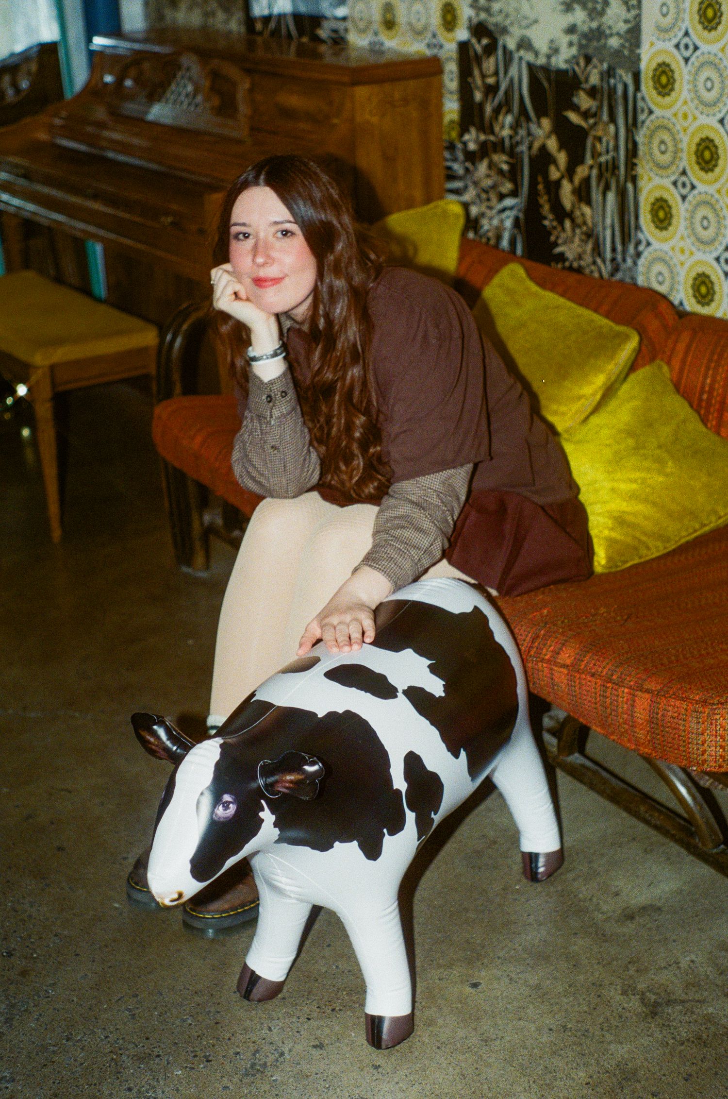 Young woman with long brown curly hair sits on decorative cow figurine in eclectic vintage room.