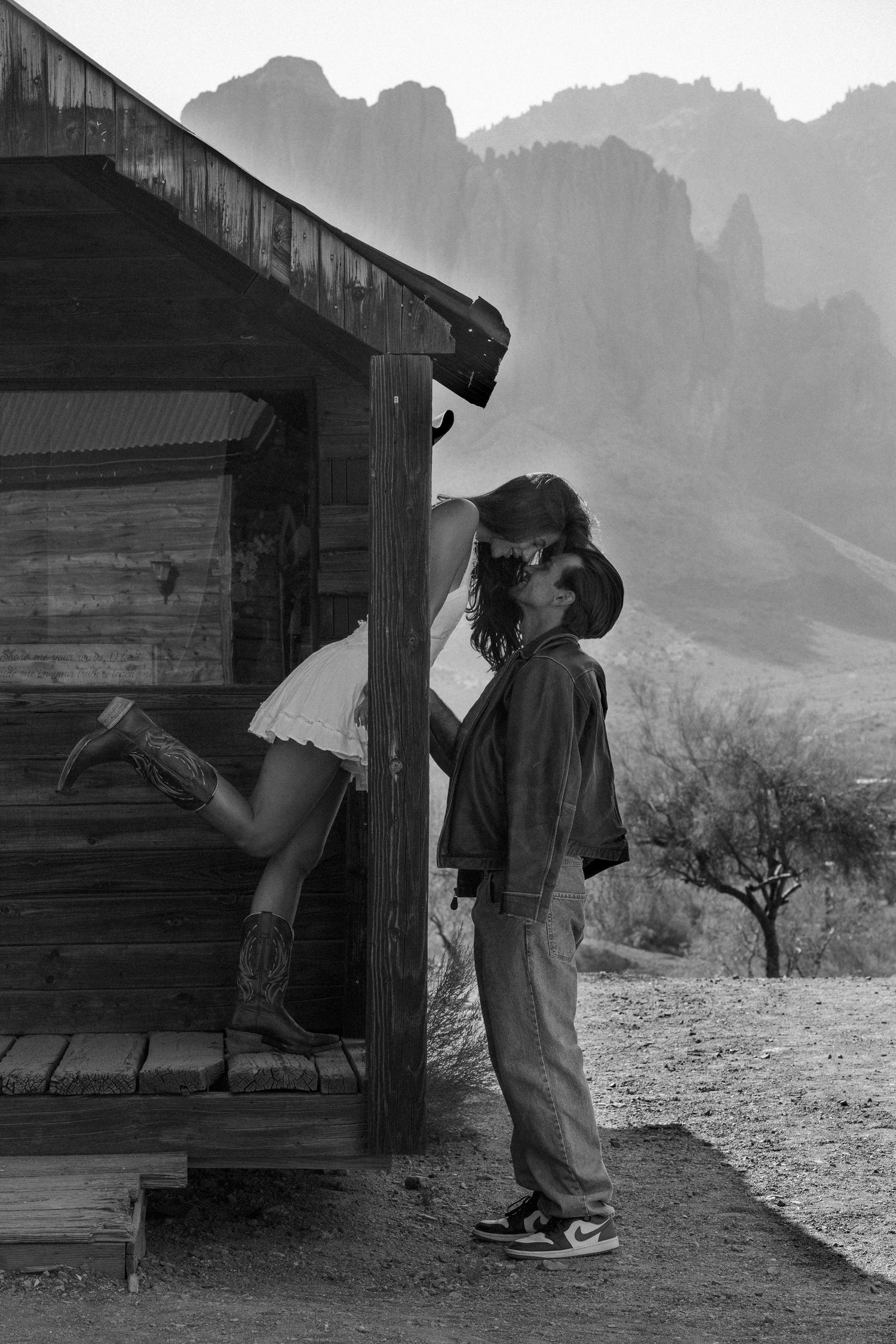 Couple sharing a kiss outside rustic wooden cabin with mountain backdrop in black and white.