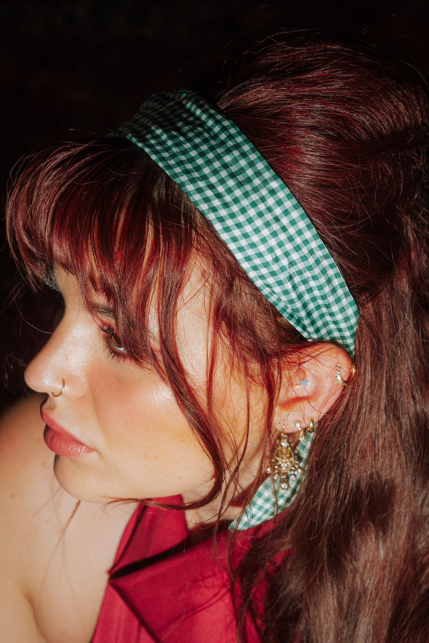 Close-up profile of a woman with red hair wearing a green gingham headband and gold ear cuff earrings.