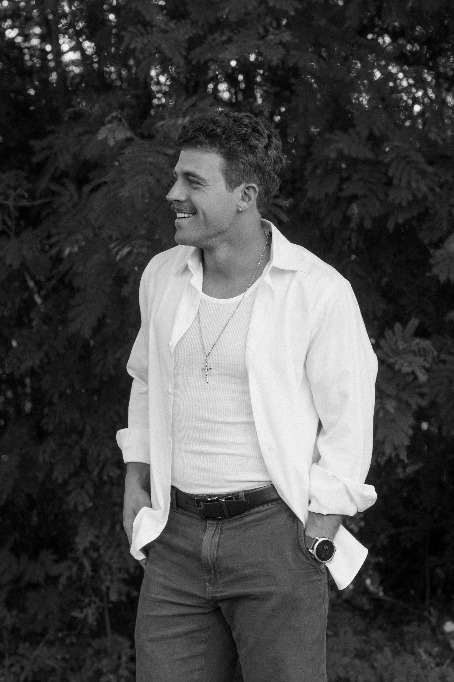Black and white portrait of smiling man in white shirt with cross necklace outdoors.