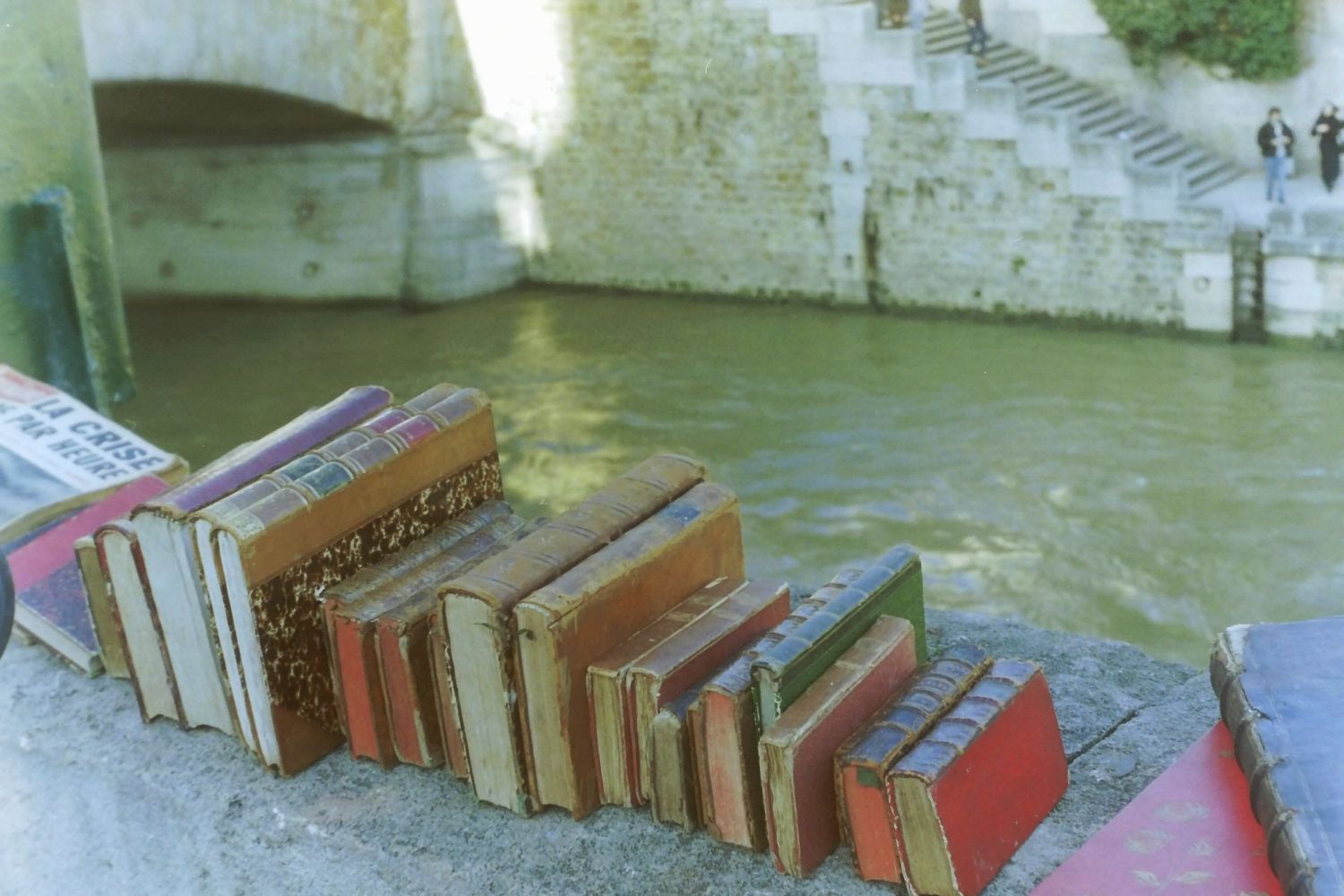 Vintage books arranged in a staircase pattern on stone steps beside a green river with a bridge backdrop.