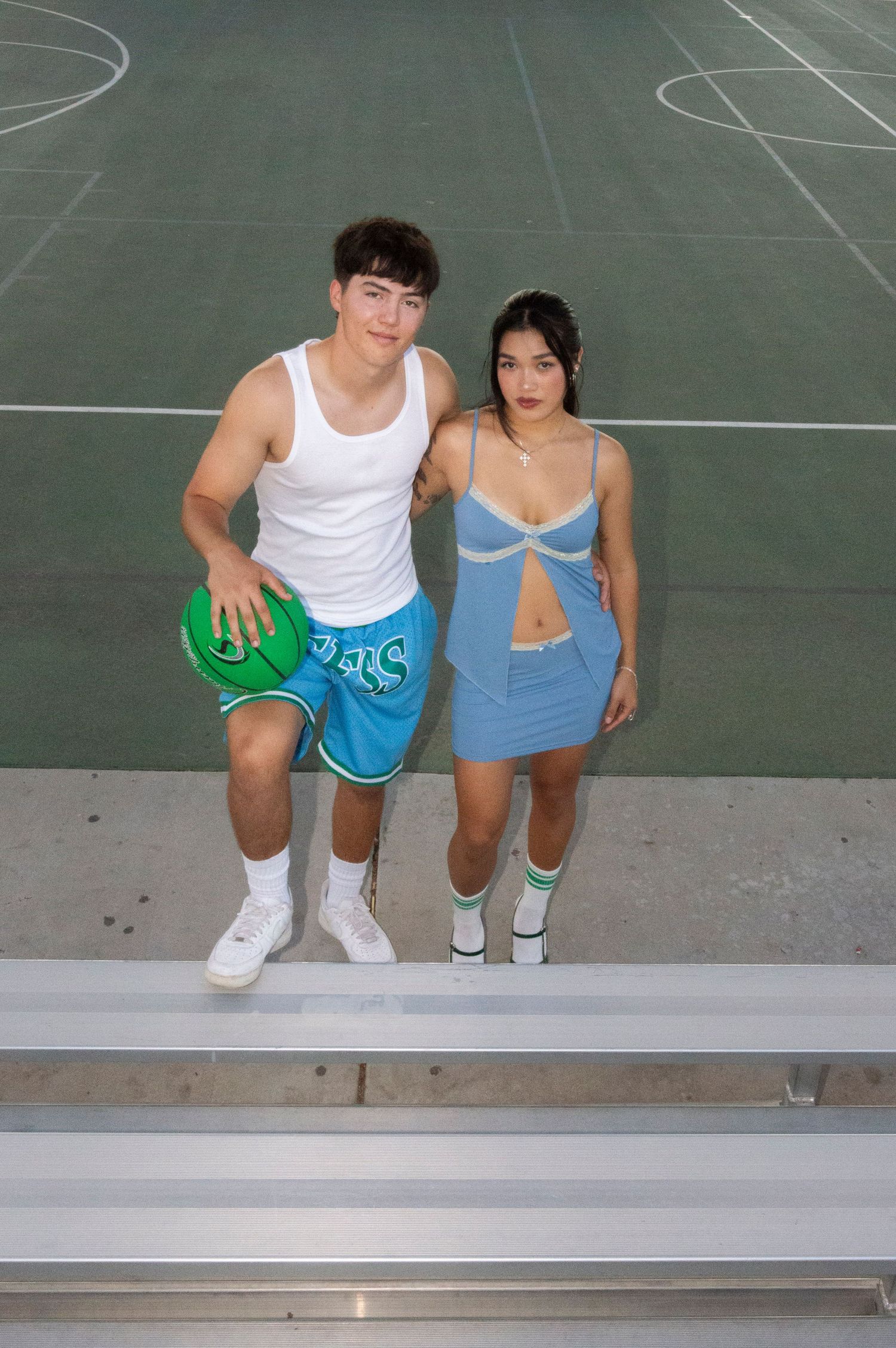 Young couple posing on bleachers at basketball court, man holding green ball, woman in blue dress.