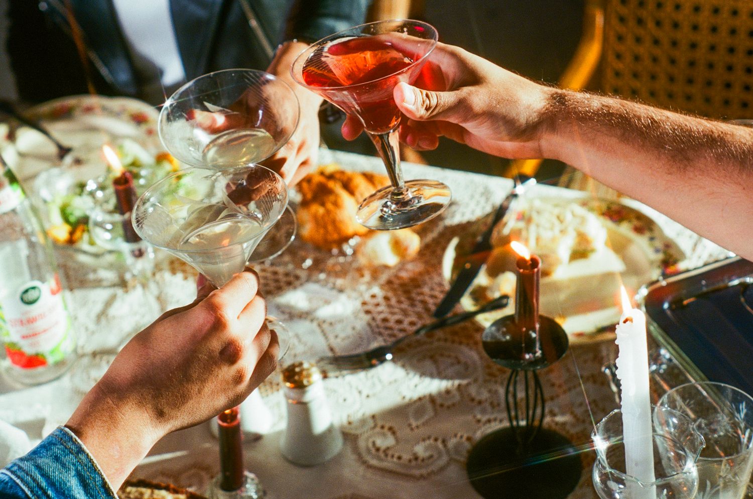 Friends toasting cocktail glasses over a candlelit dinner table.