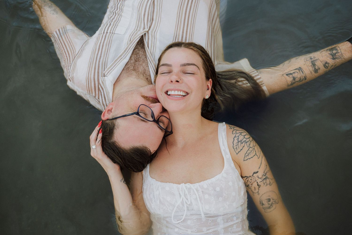 Couple lying on ground smiling, woman with tattoos in white dress, man with glasses kissing her cheek.