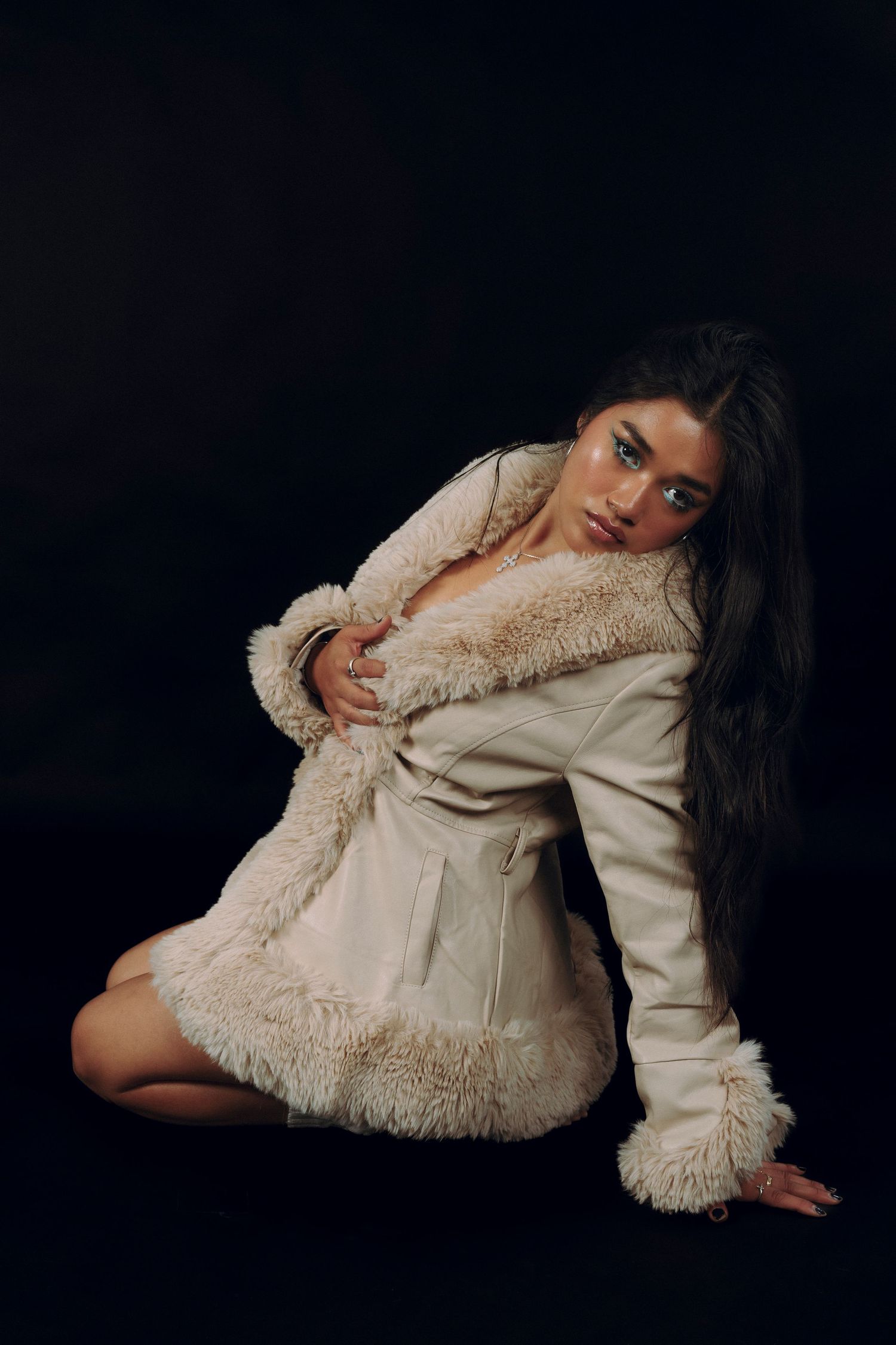 Woman in cream shearling coat posing on floor against dark background in fashion editorial style photo.