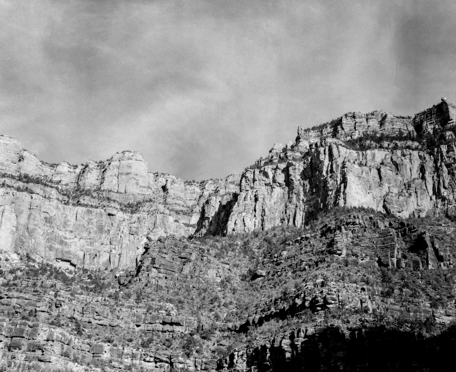 Black and white photo of dramatic rocky canyon cliffs with layered geological formations under a cloudy sky.