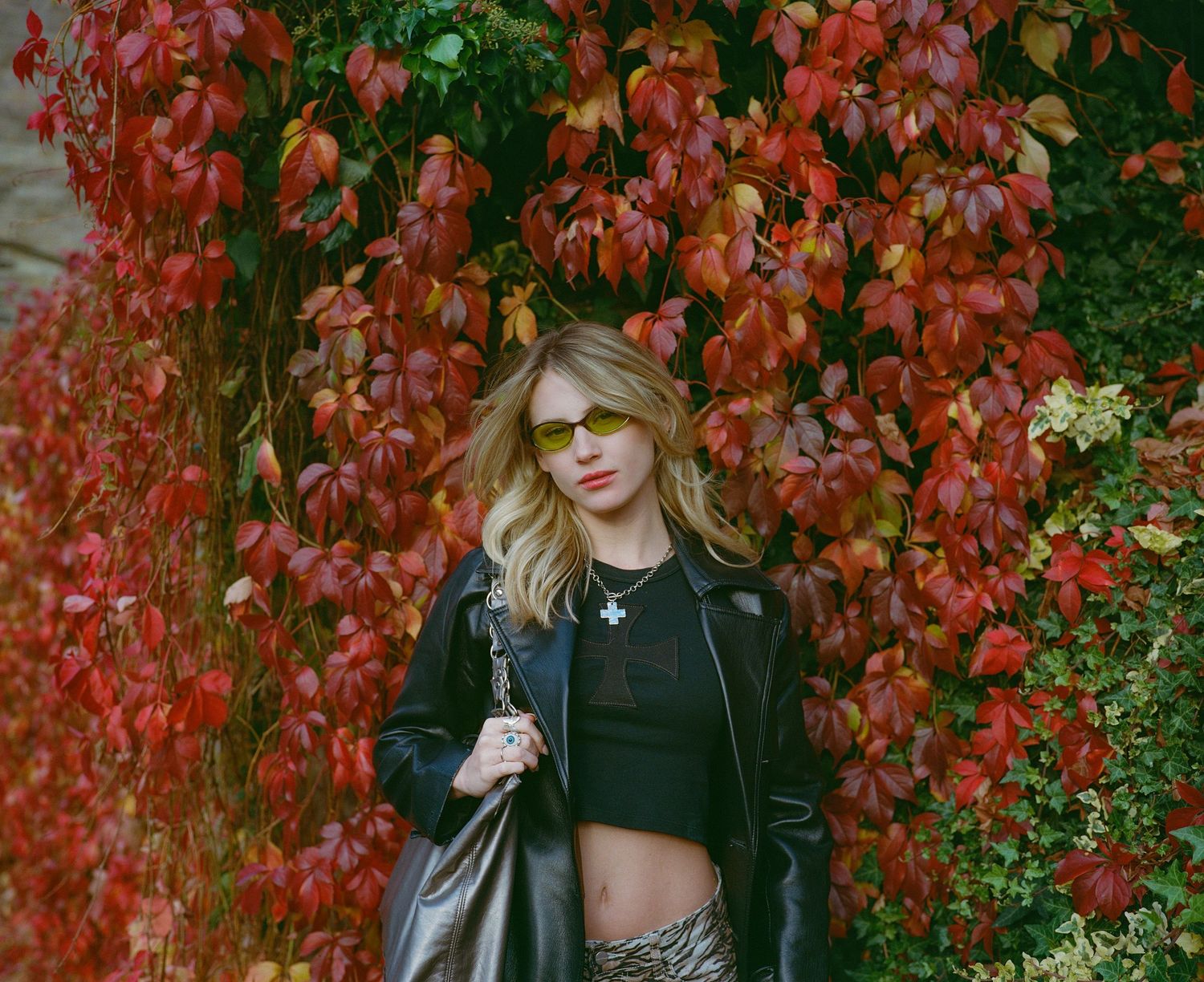Blonde woman in black leather jacket and crop top posing against vibrant red autumn ivy wall.