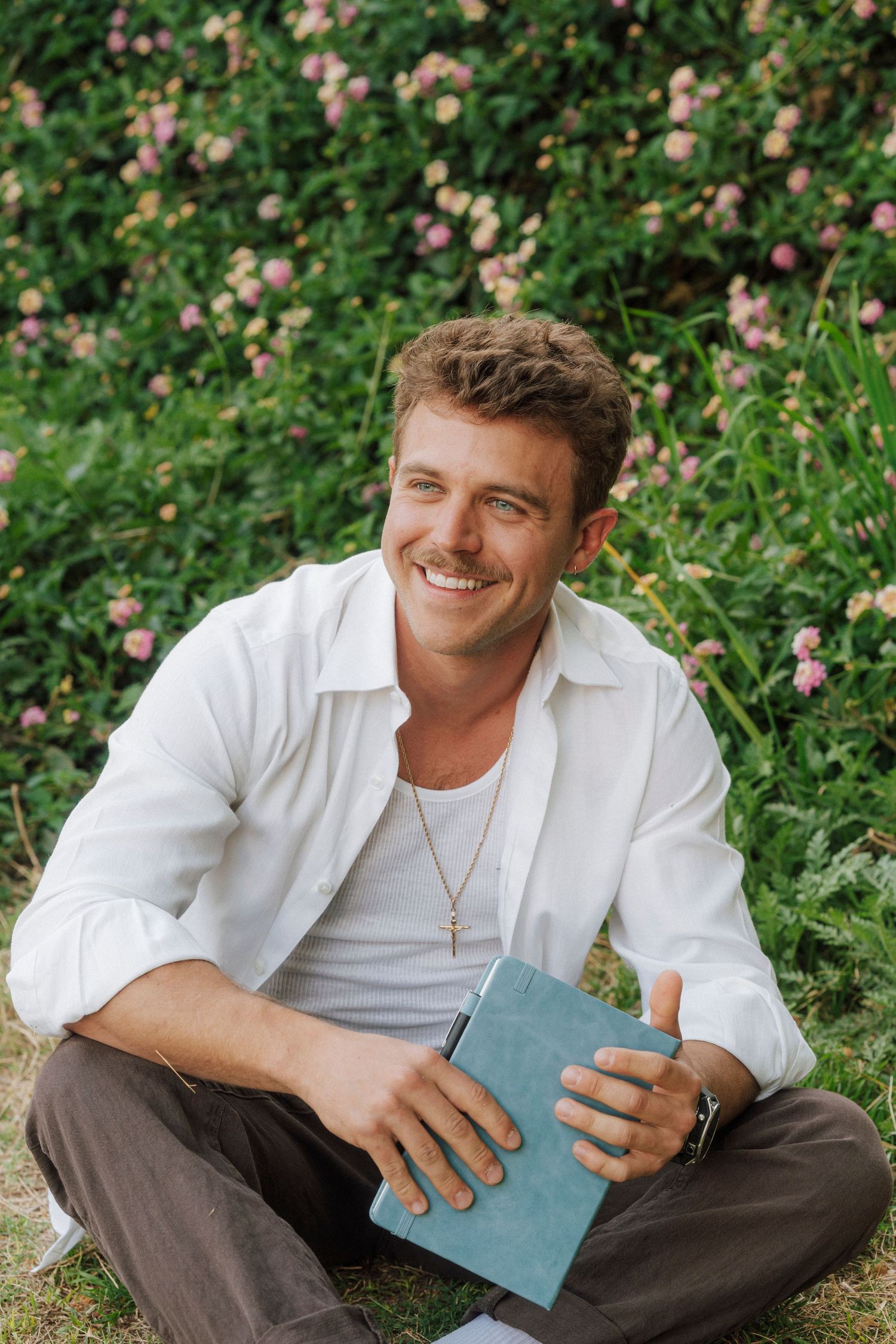 Smiling man in white shirt holding a blue book, sitting cross-legged outdoors near pink flowering bushes.