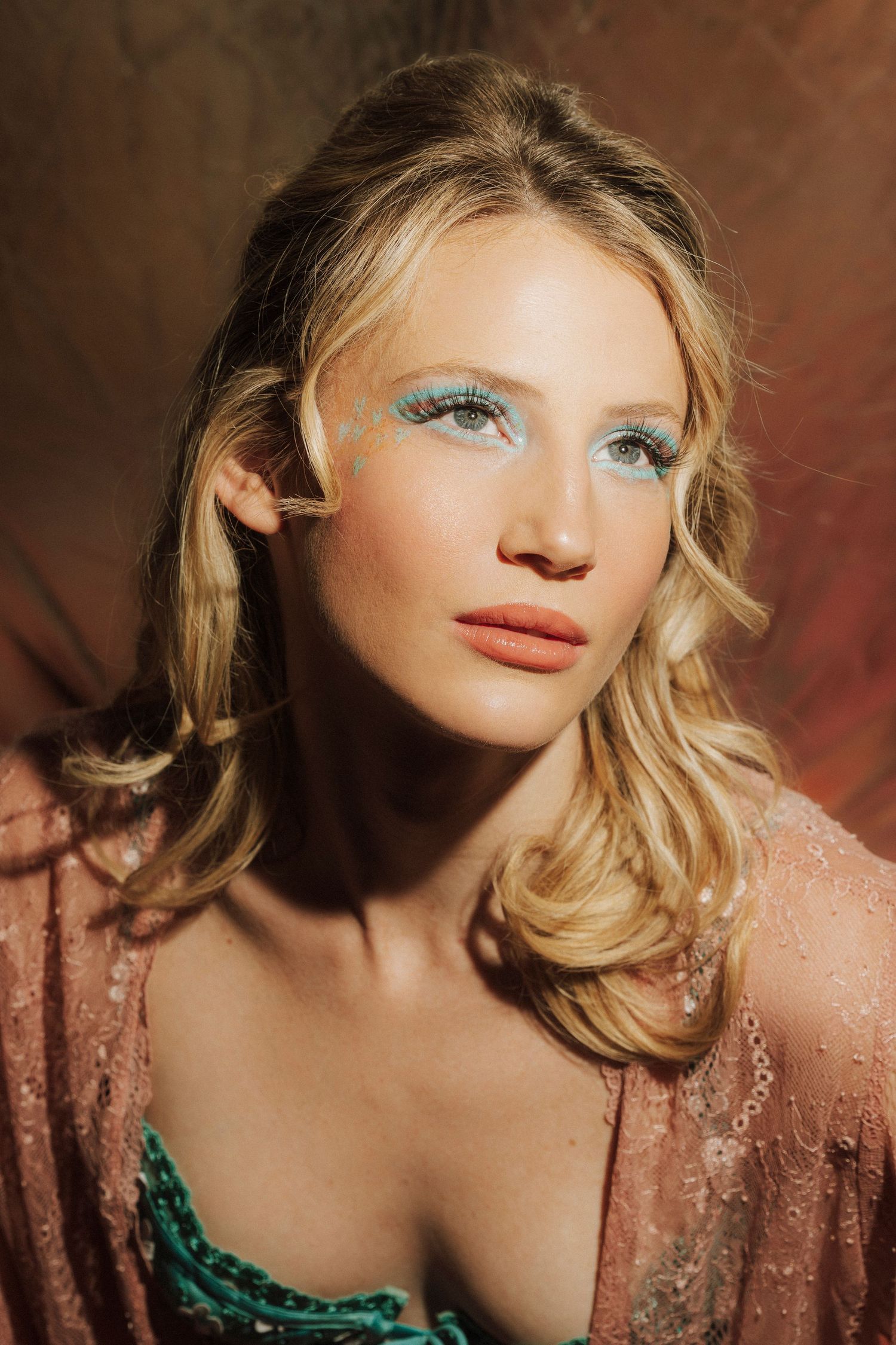 Blonde woman with teal artistic eye makeup in pink sheer top in warm dramatic lighting.