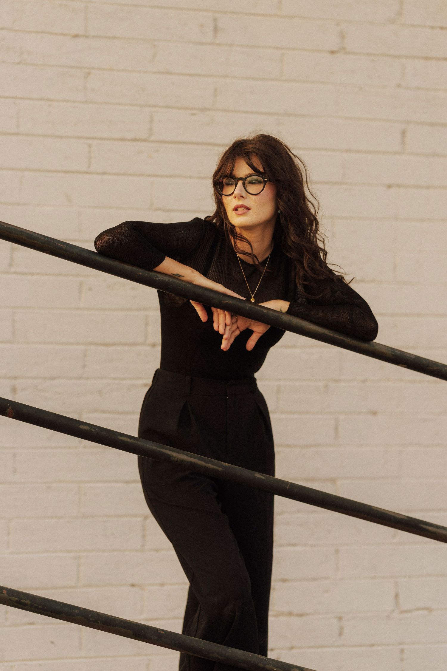 Woman in black outfit and glasses leaning on metal railing against white brick wall.