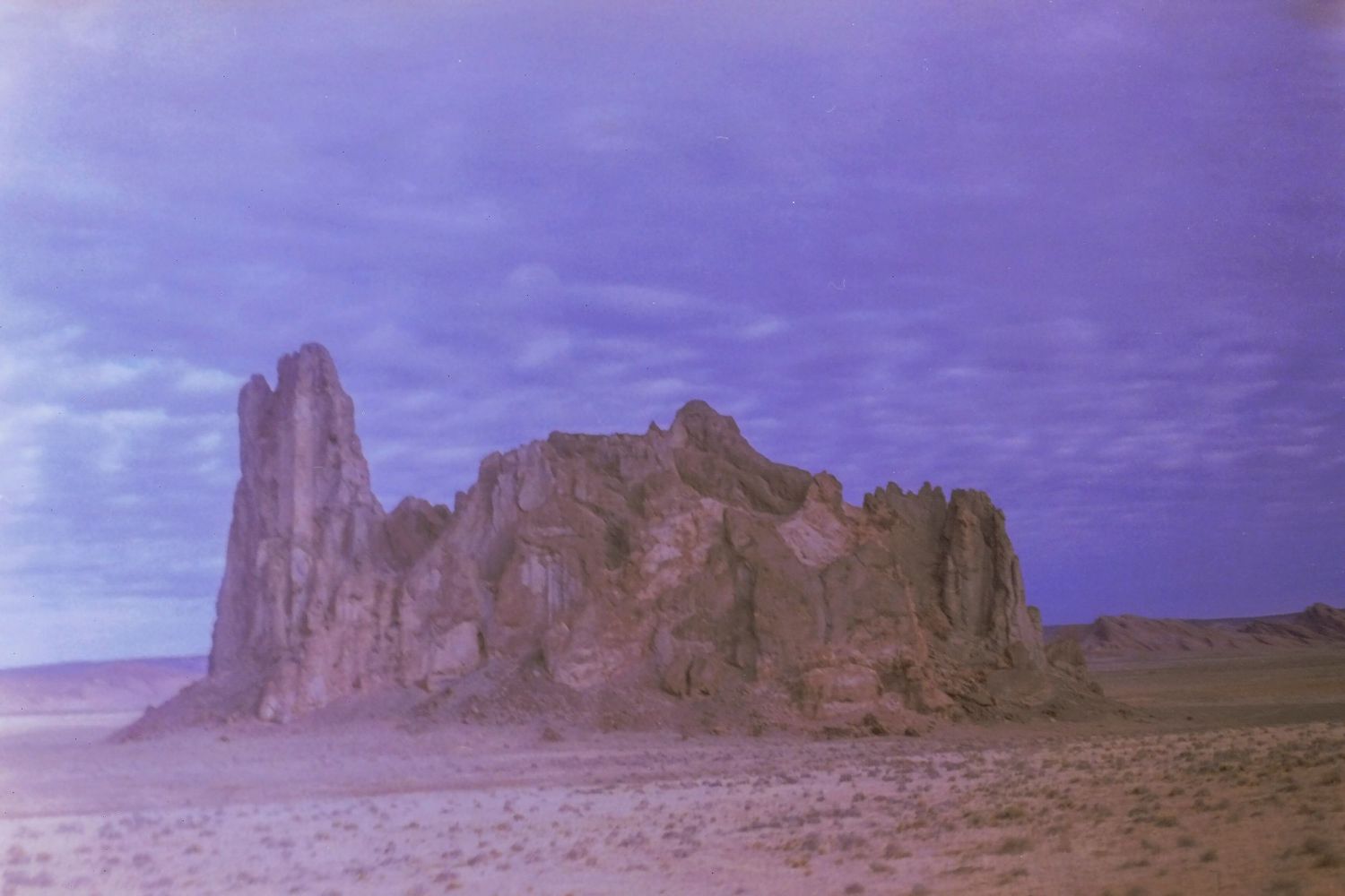 Purple-tinted desert landscape with dramatic rock formations under cloudy sky.