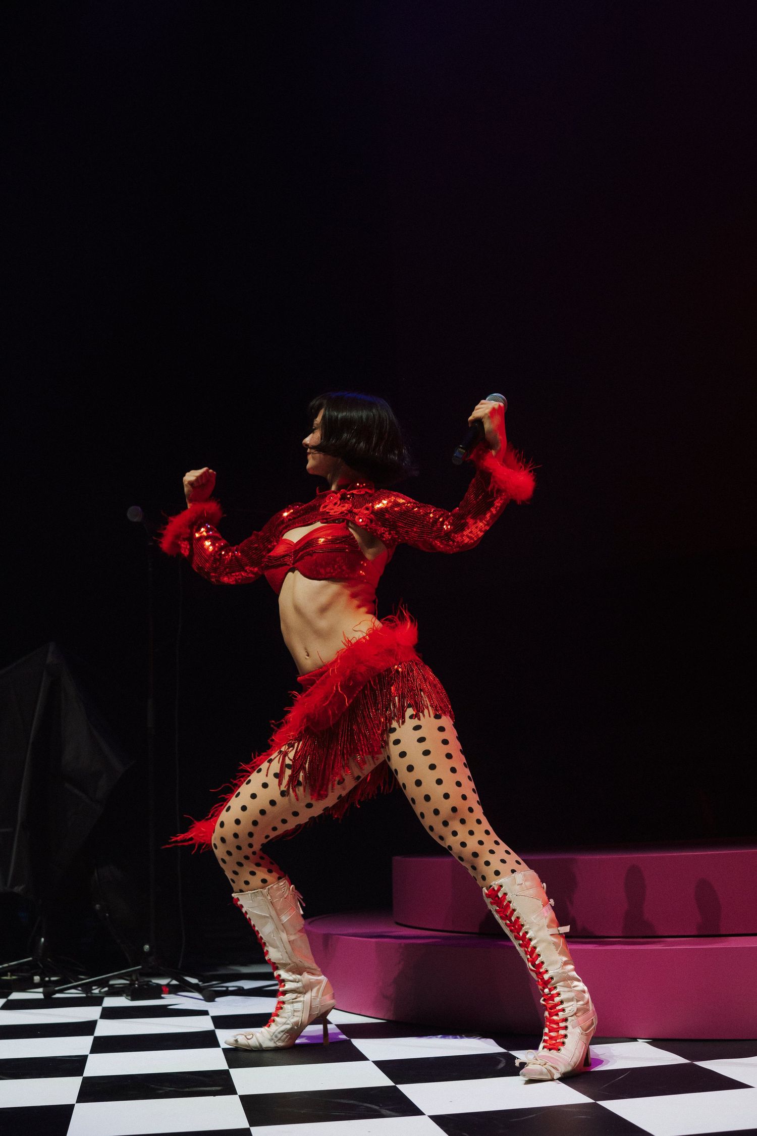 Performer in red sequined crop top, polka dot tights, and white boots dancing on checkered floor stage.