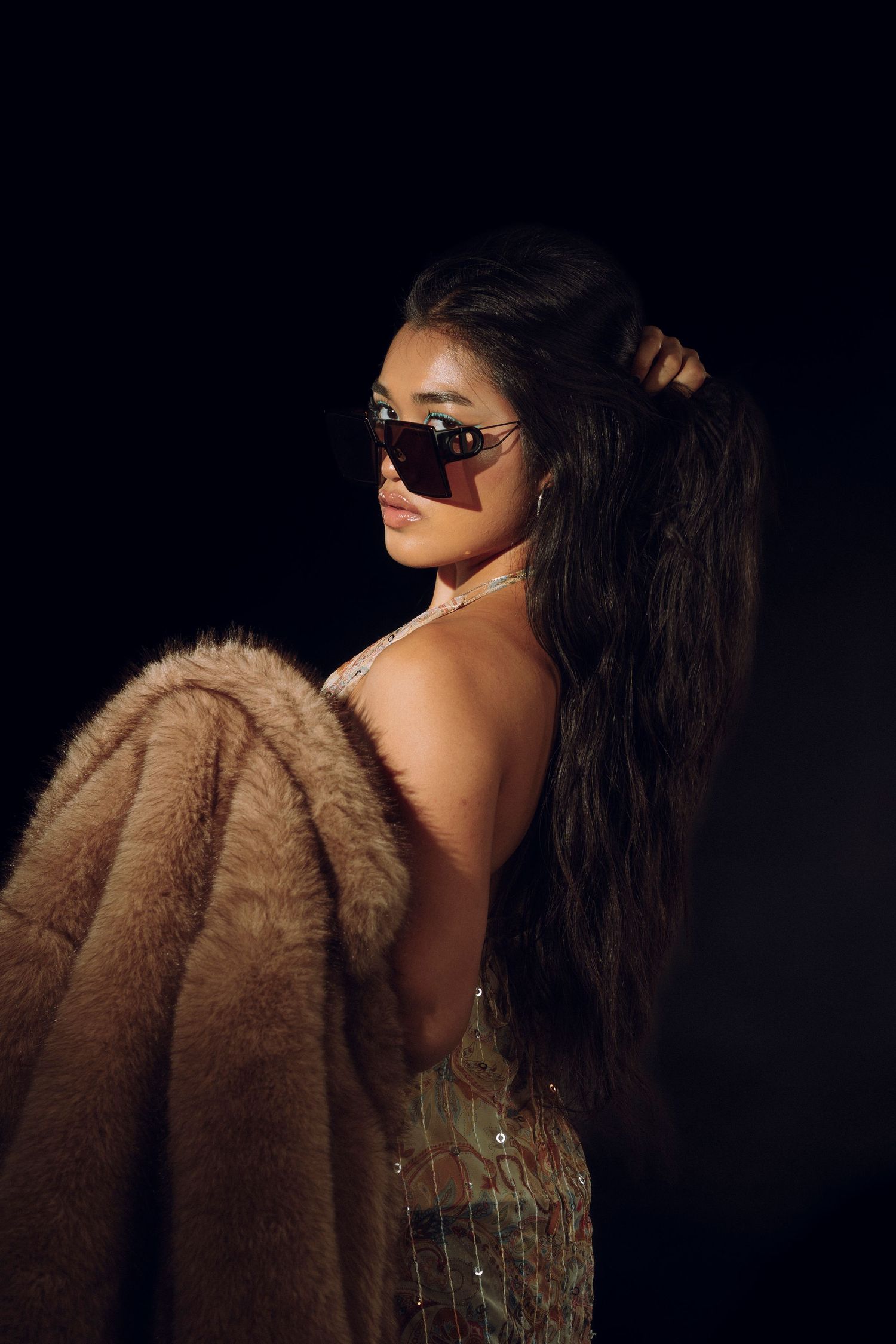 Woman in faux fur coat and oversized sunglasses poses against dark background with long flowing hair.